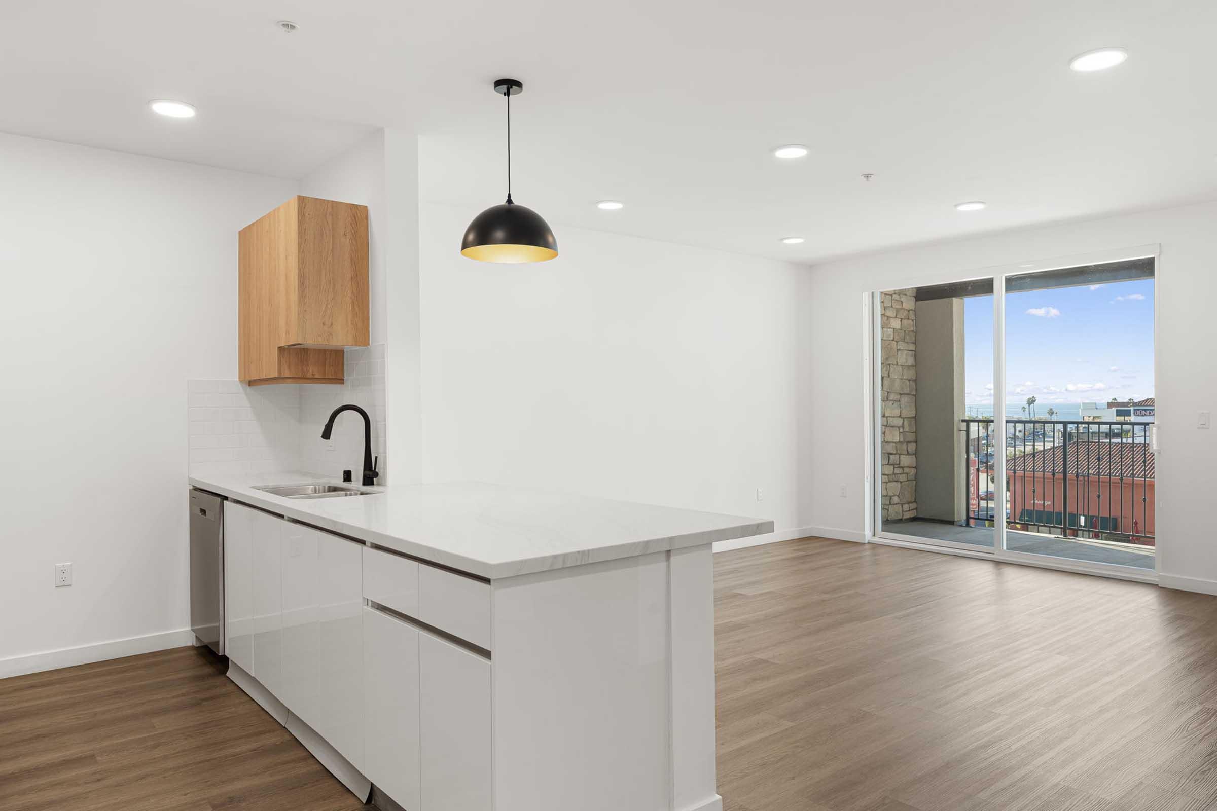 Modern kitchen and living area in a bright apartment. The kitchen features a white countertop and sleek cabinetry, with a black pendant light above the island. Large sliding glass doors lead to a balcony, revealing a view of the outdoors. The flooring is light wood, enhancing the spacious feel.