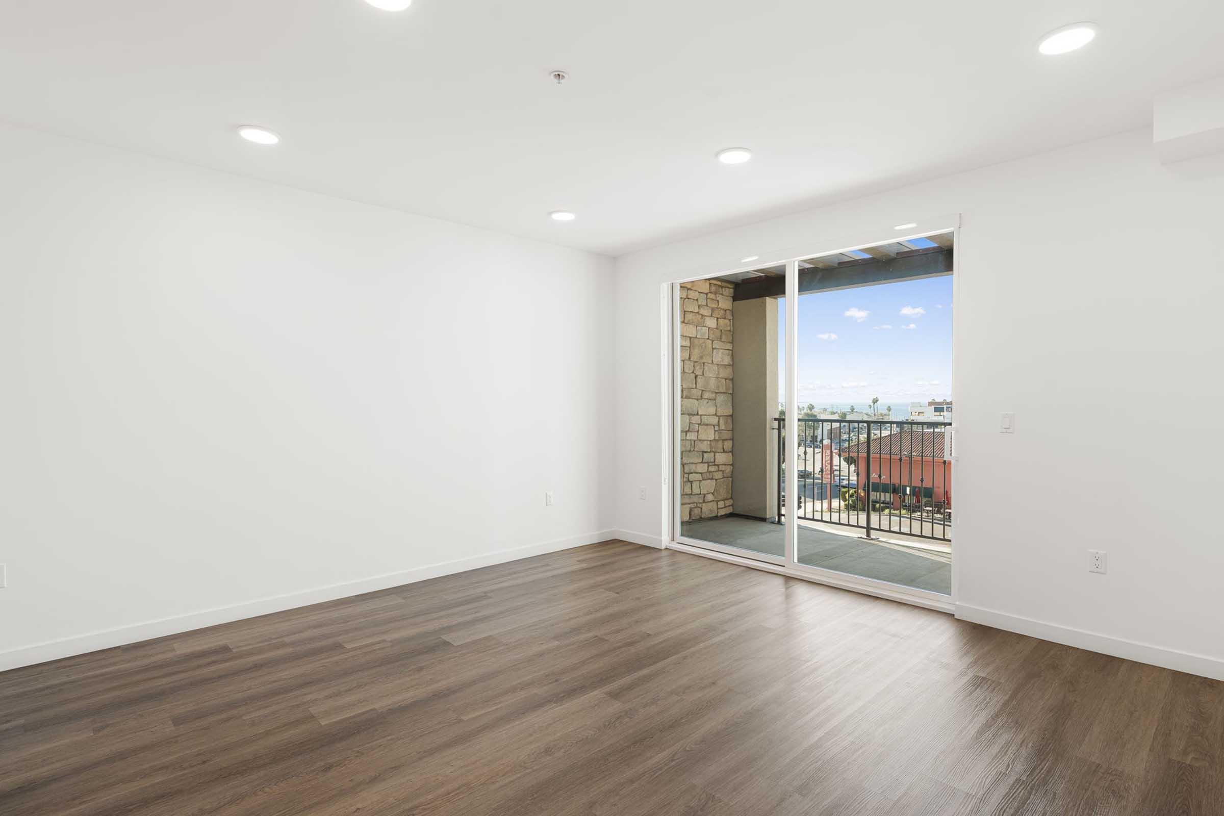 Empty room with a wooden floor, white walls, and recessed lighting. A large sliding glass door opens to a balcony with a view of the outdoors. The space is bright and airy, suggesting a contemporary design.