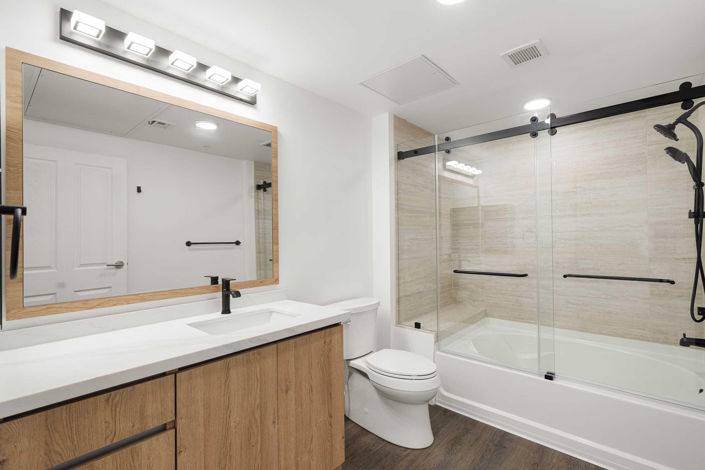 Modern bathroom featuring a rectangular mirror with LED lighting, a sleek vanity with wooden cabinetry, a white sink, a toilet, and a glass shower enclosure with black hardware. The walls are light-colored, and the floor has a wooden finish, creating a clean and contemporary look.