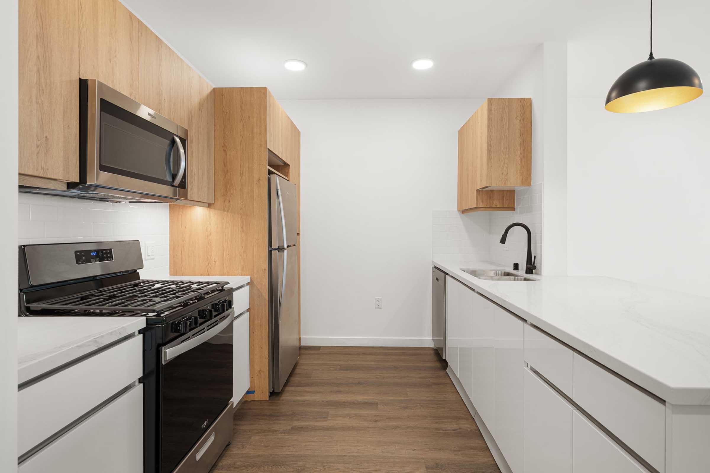 Modern kitchen featuring stainless steel appliances, including a gas range and refrigerator. The cabinetry is a mix of white and light wood, with a clean, minimalist design. Bright overhead lights illuminate the space, highlighting the sleek countertop and sink. A pendant light hangs above the kitchen island for added style.