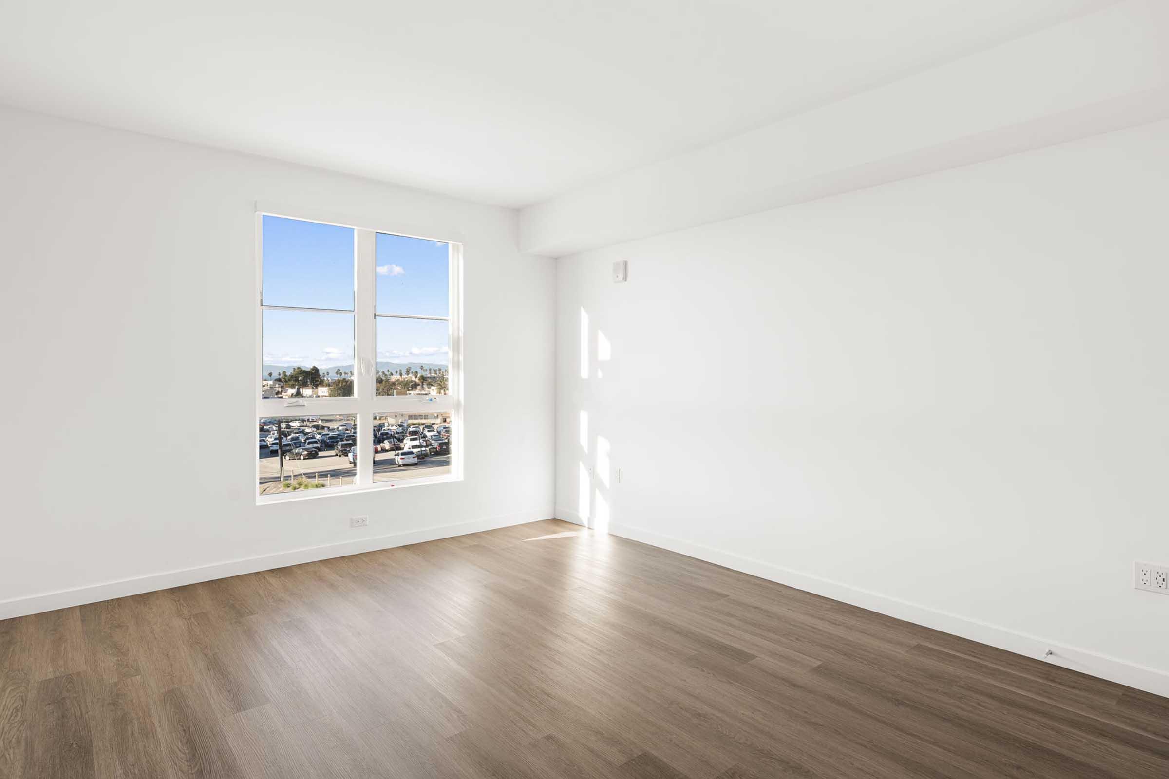 A bright, empty room with light wood flooring and white walls. A large window allows natural light to fill the space, providing a view of the outdoors. The overall atmosphere is spacious and modern, ideal for customization and personal decoration.
