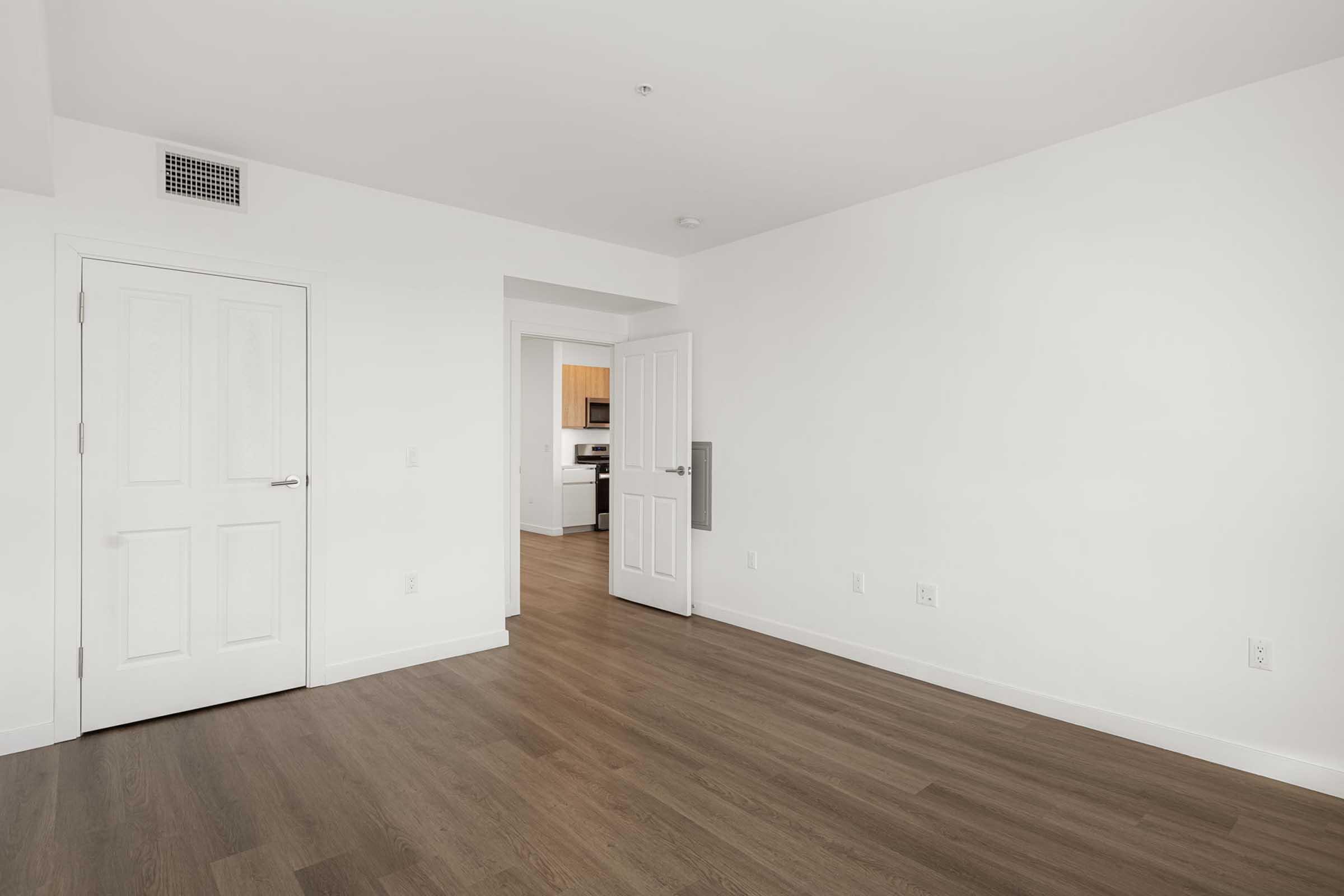 Empty room featuring light hardwood floors and white walls. A closed door on the left leads to another space, while an open doorway on the right gives a view of a kitchen area. Natural light filters in, creating a bright and spacious atmosphere.