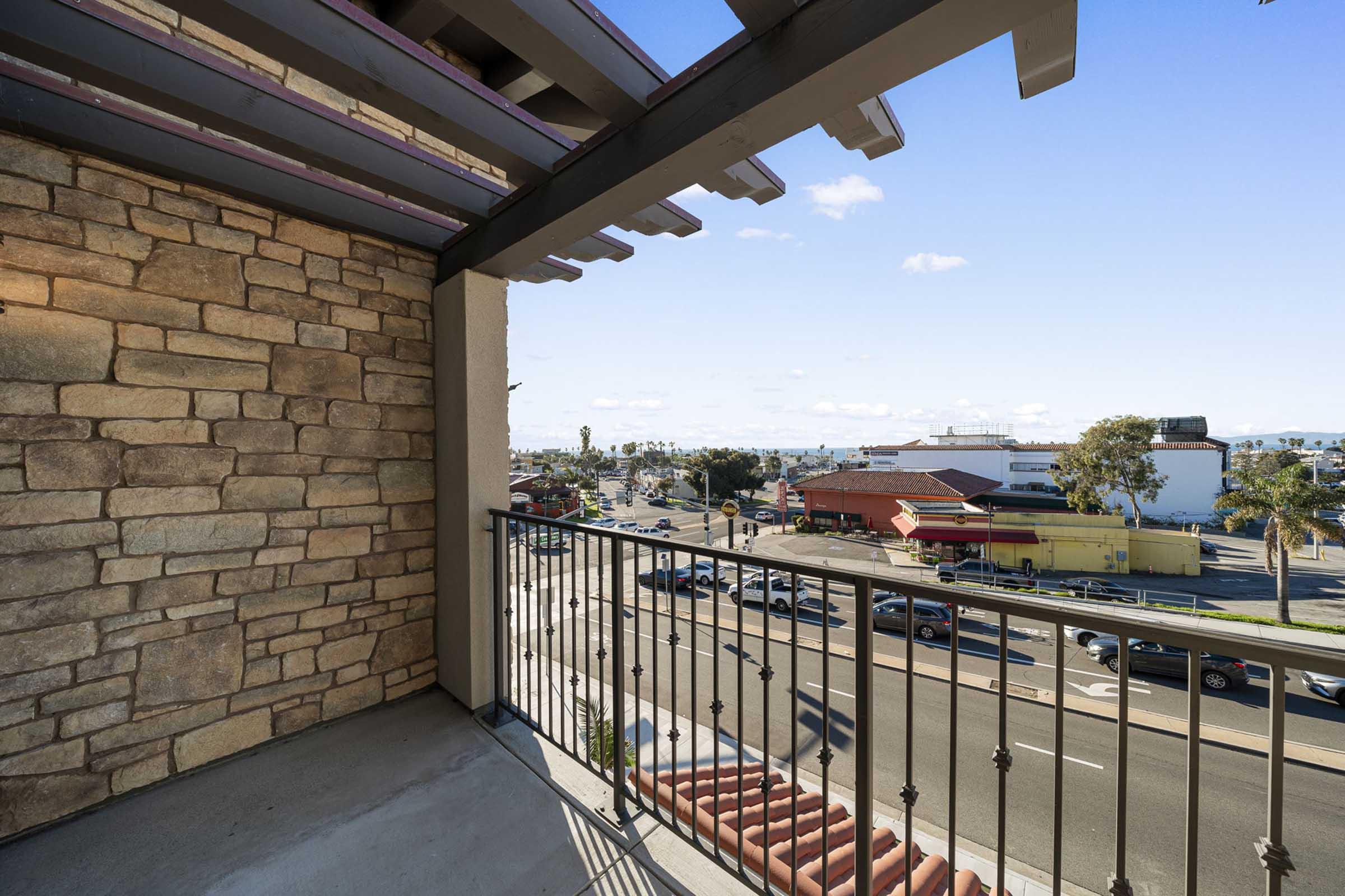 View from a balcony showcasing a cityscape with a mix of buildings, including restaurants and shops, set against a clear blue sky with scattered clouds. The balcony features a stone wall and a railing, providing an open perspective of the bustling street below.