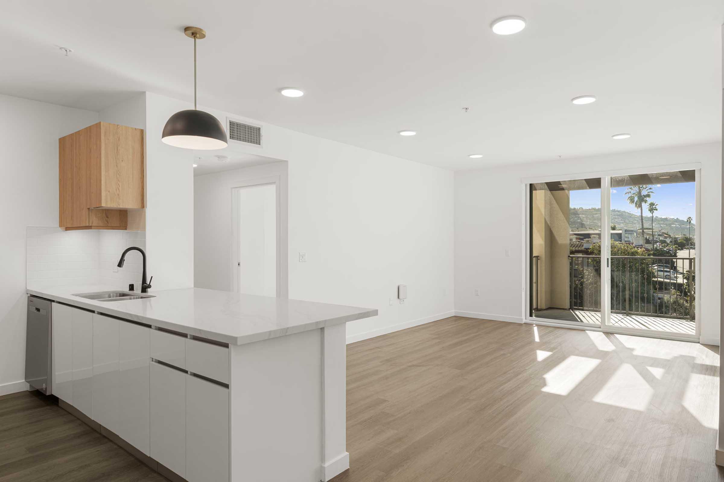 A modern, bright apartment interior featuring a spacious kitchen with white cabinetry and a wooden accent, a living area with large windows, and sliding glass doors leading to a balcony. The floor is light wood, and the walls are painted white, creating a clean and airy atmosphere.