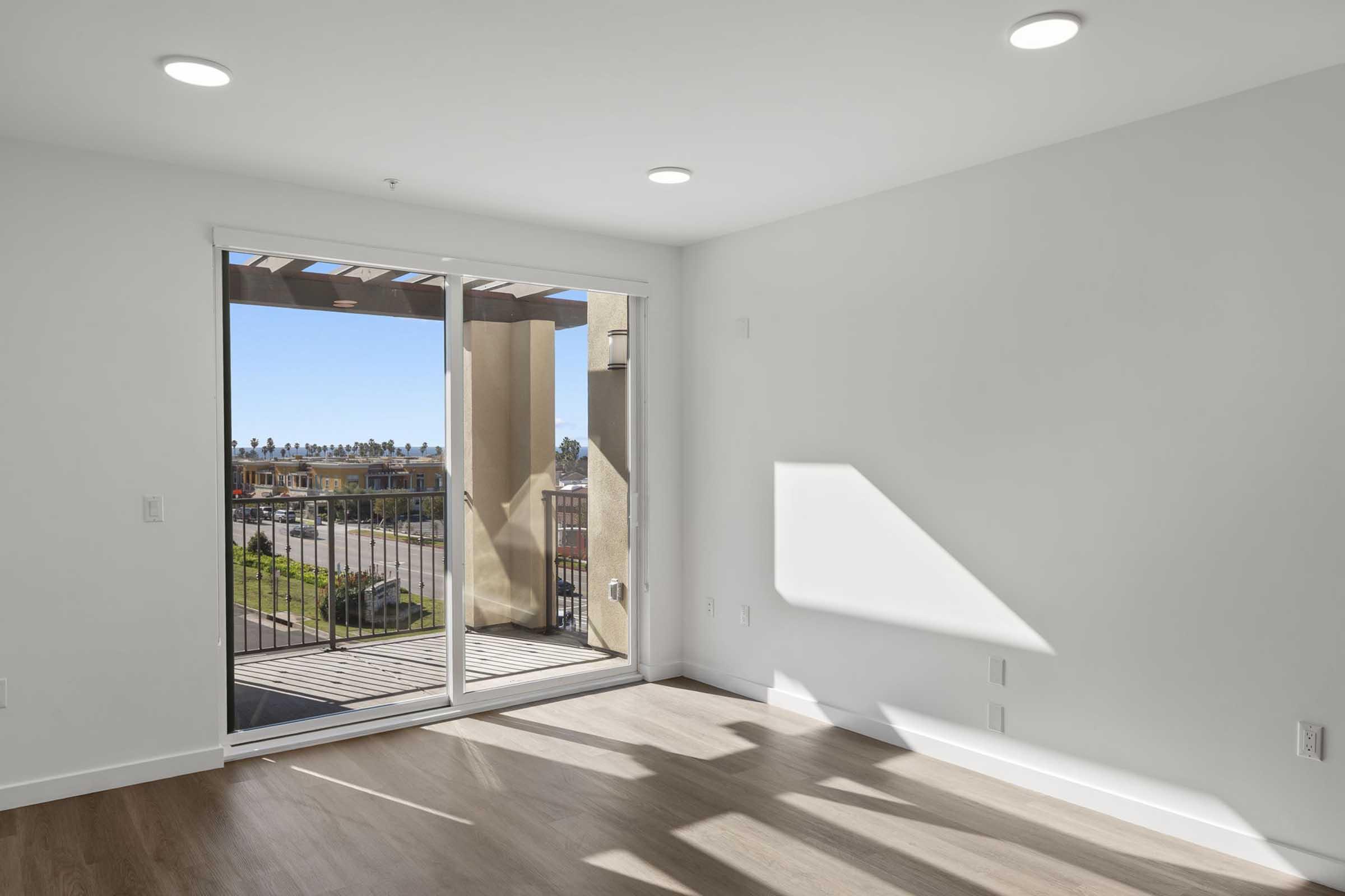 Bright, empty room with light wood flooring and white walls. A large sliding glass door leads to a balcony with a view of a grassy area and distant buildings. Natural light streams in from the windows, creating a welcoming atmosphere.
