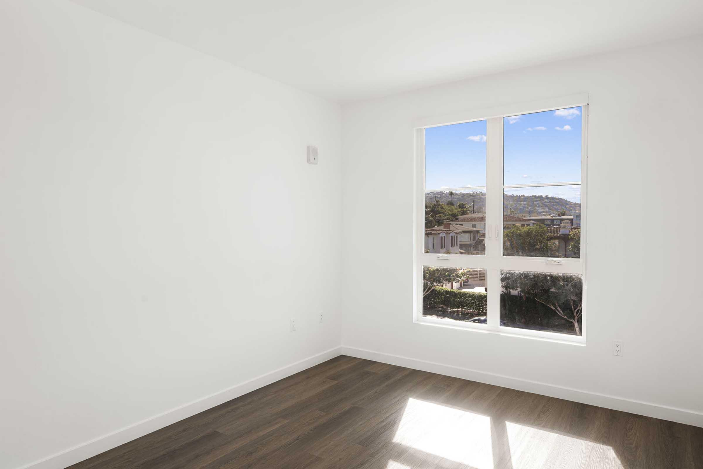 A bright, empty room with white walls and a large window letting in natural light. The window offers a view of a hilly landscape and blue sky, while the floor is polished dark wood.