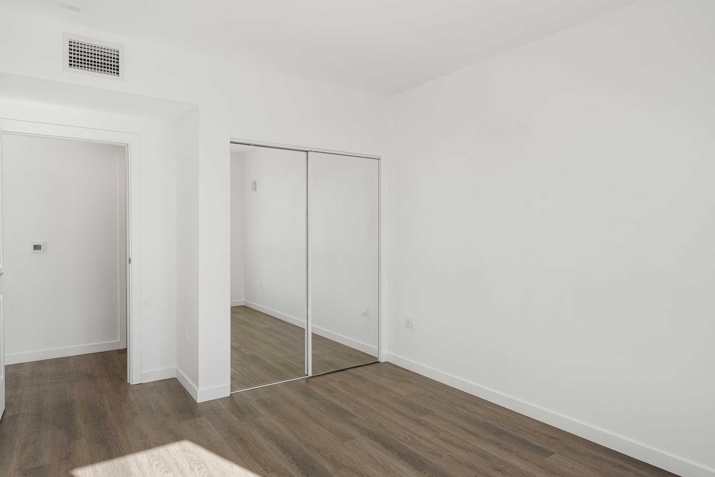 A minimalistic interior room featuring light-colored walls and a wooden floor. There is a doorway on the left leading to another space, and a mirrored closet door reflects the empty room. The area is bright and uncluttered, creating an open and airy atmosphere.