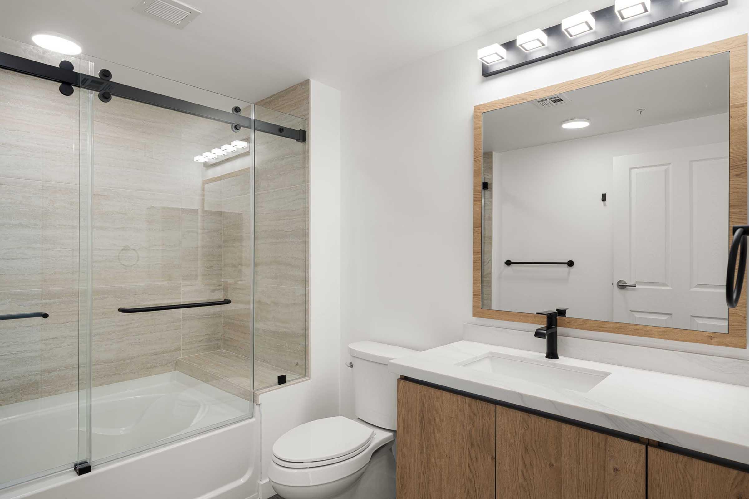 Modern bathroom featuring a glass shower enclosure, a white bathtub, contemporary sink with a faucet, wooden vanity with storage, large mirror, and stylish lighting. Neutral tones and sleek design elements create a clean and spacious feel.