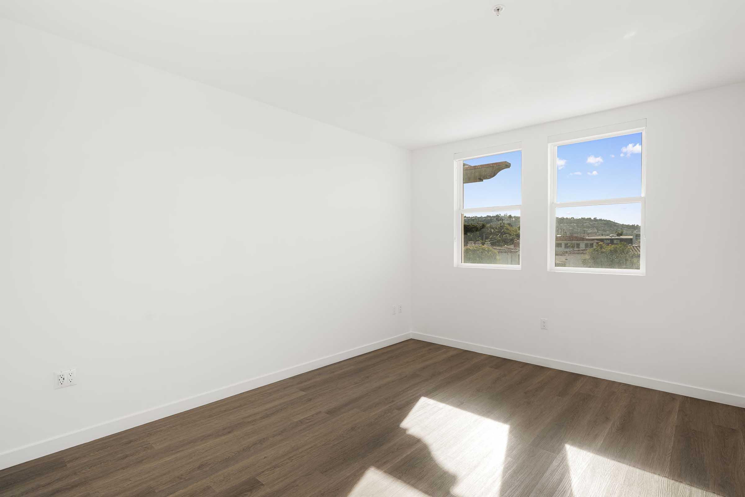 Spacious empty room with light-colored walls and large windows allowing natural light. Wooden flooring adds warmth, while a scenic view is visible through the windows. The room is ready for furnishing and personalization.