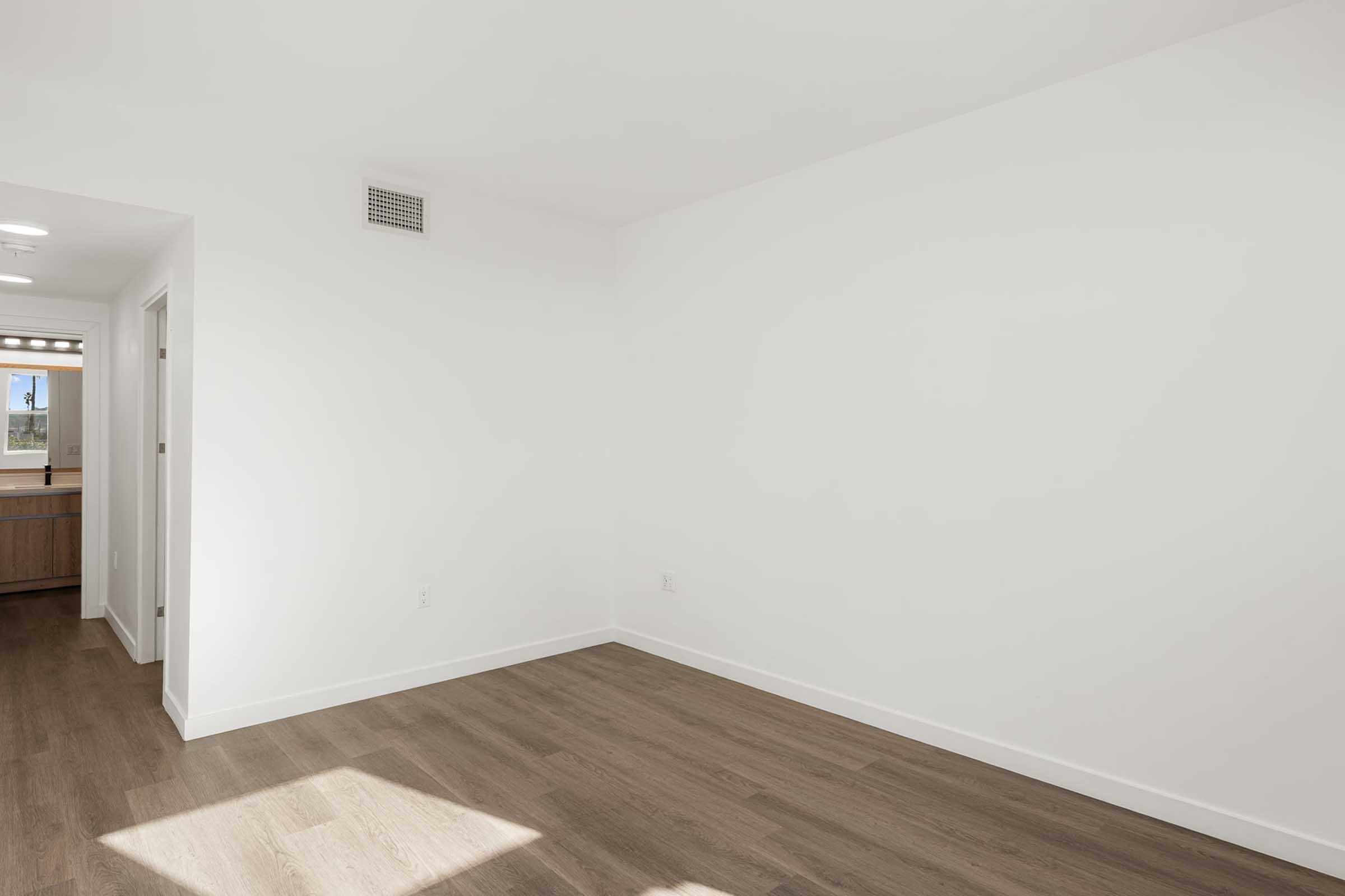 A bright, empty room with white walls and a large window allowing natural light. The floor is made of wooden planks, and there is a doorway leading to another room with wooden cabinetry visible in the background. The space has a minimalist aesthetic and is ready for furnishing.