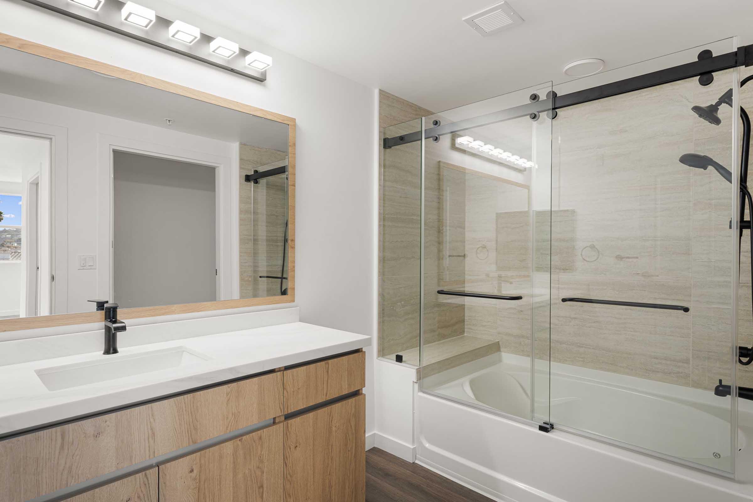 A modern bathroom featuring a glass shower enclosure, a white bathtub, a large mirror with wooden framing, and a sleek vanity with a dark faucet. The walls are tiled with light-colored stone, and there is a bright, well-lit atmosphere with natural light visible from a nearby window.