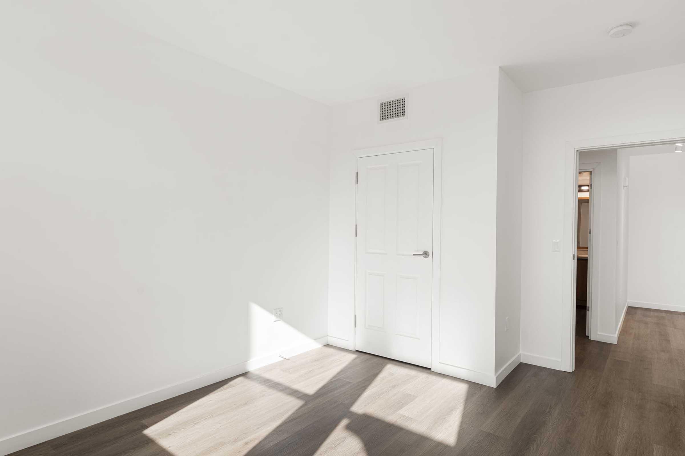 A bright, empty room with white walls and light wood flooring. A closed white door is visible on the left, with sunlight streaming in from a nearby window, casting shadows on the floor. An open doorway leads to another room in the background. The space is clean and minimalistic.