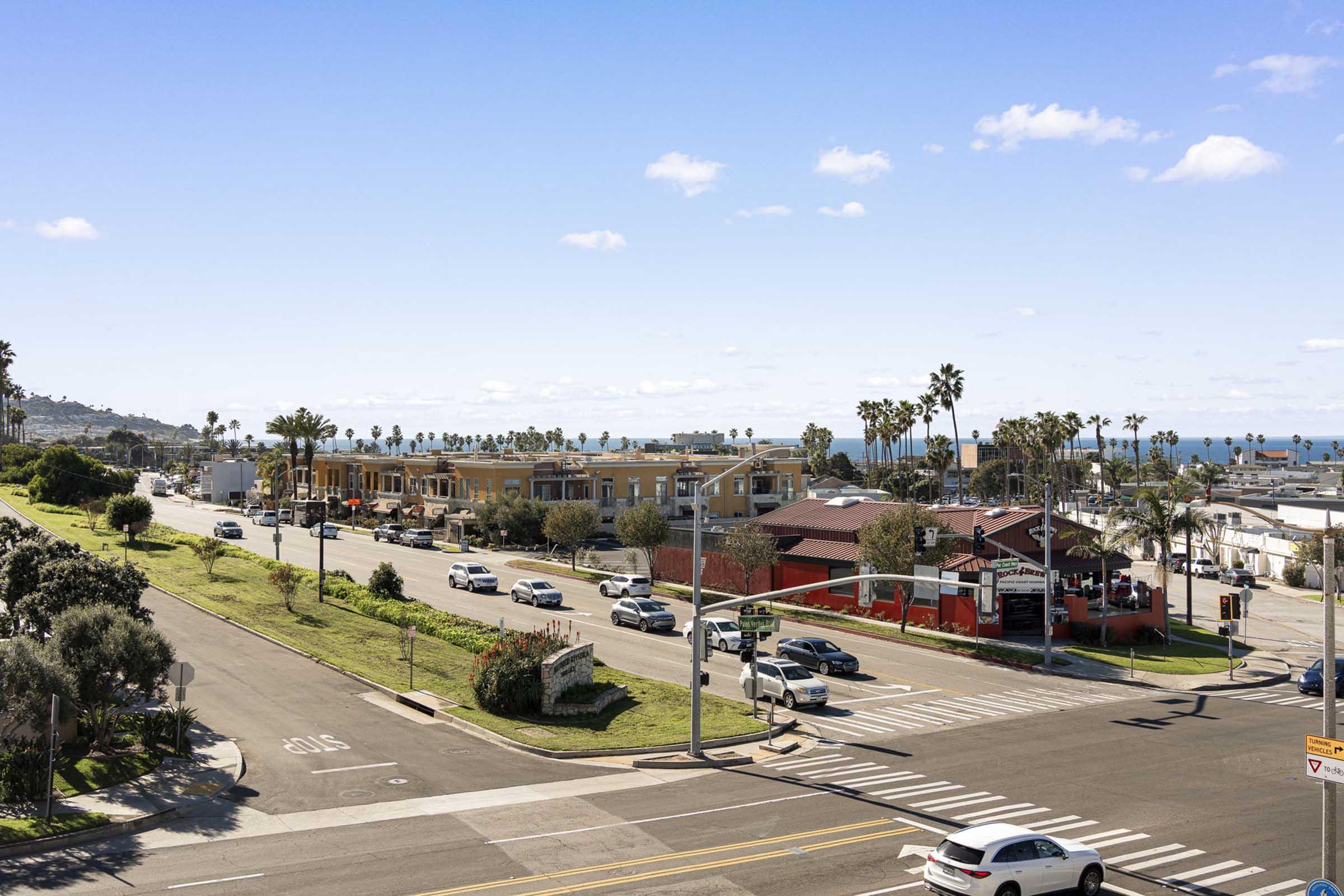 A sunny coastal street view featuring palm trees, parked and moving cars, and buildings. The ocean is visible in the background under a bright blue sky with scattered clouds. The scene captures a vibrant, relaxed atmosphere typical of a seaside town.