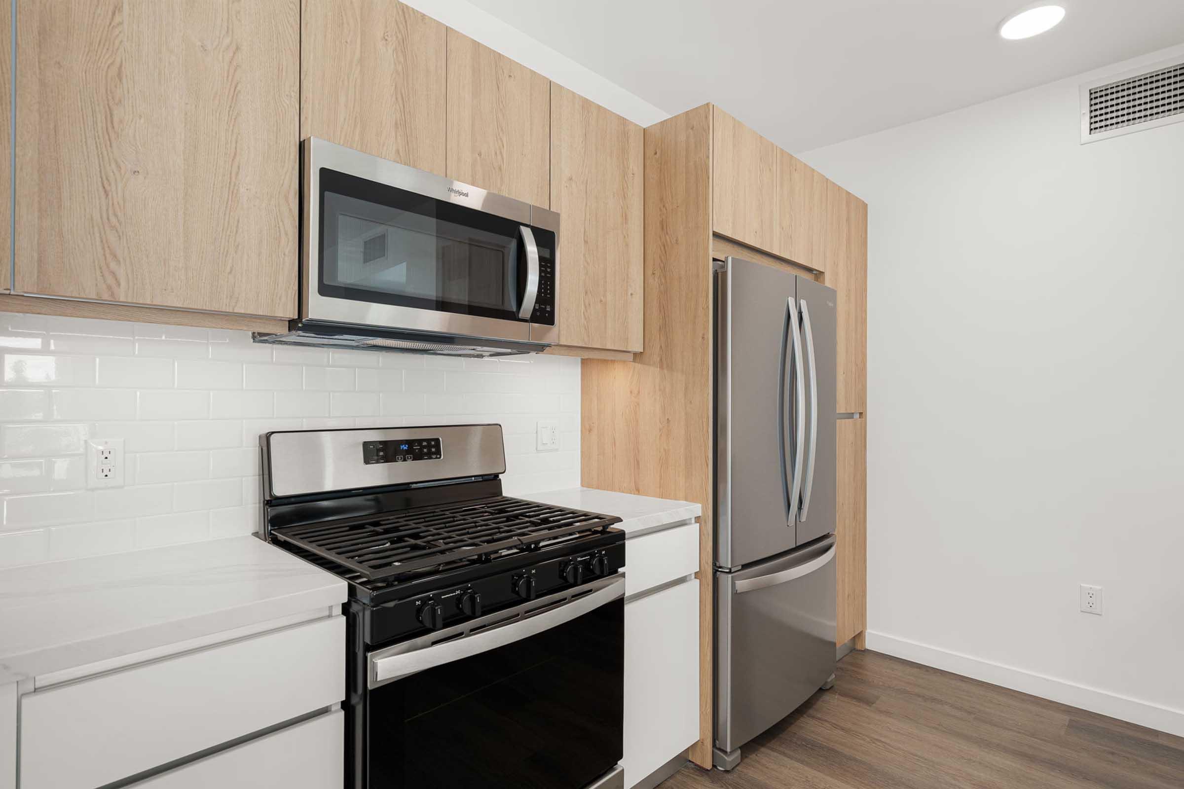 Modern kitchen featuring a stainless steel microwave, gas range with oven, and a silver refrigerator. The cabinetry is a light wood finish, complemented by a white subway tile backsplash and a sleek countertop. The flooring is a warm wood tone, enhancing the contemporary design.