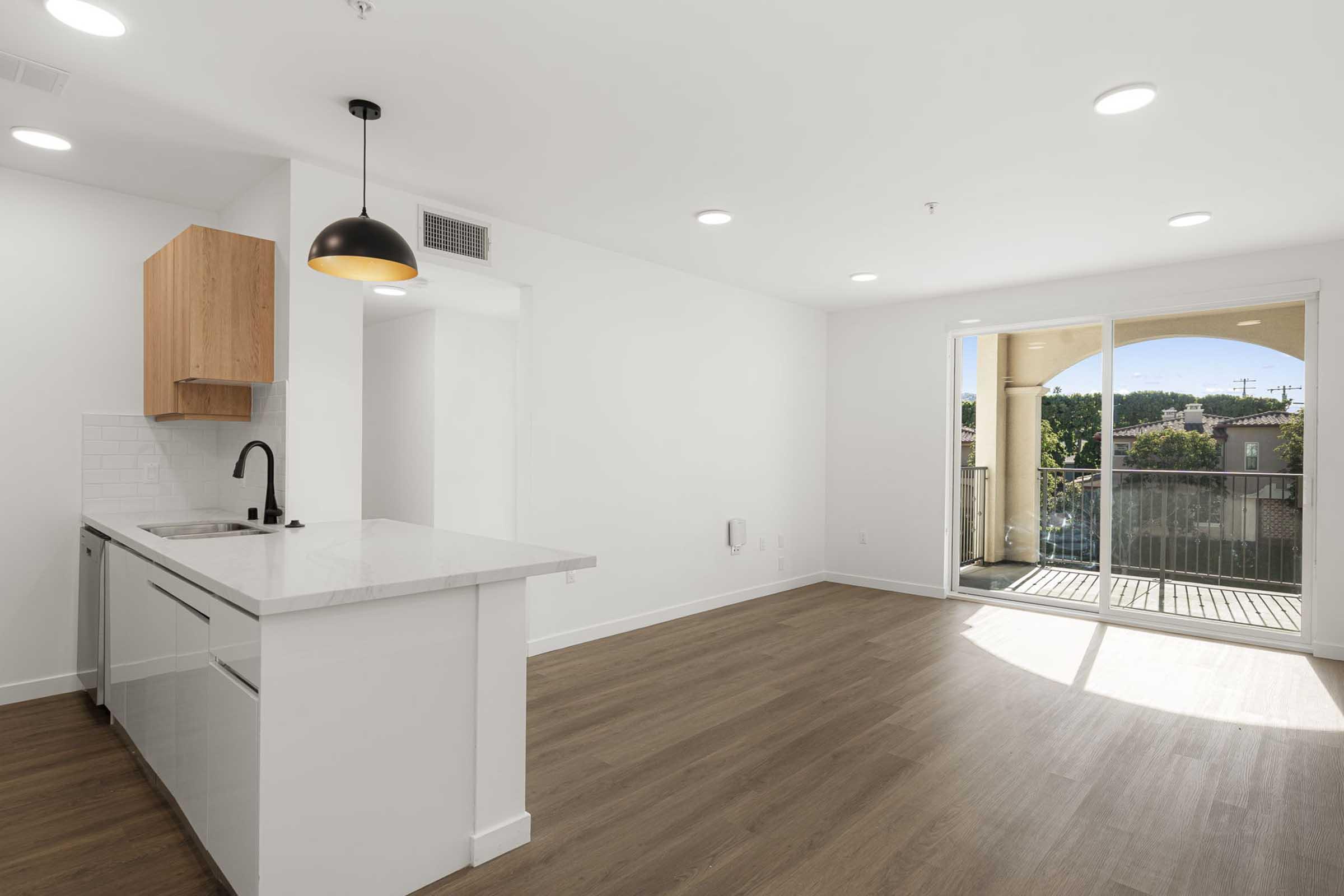 Modern, open-concept living space with a kitchen featuring a white countertop and wooden cabinetry. There’s a sink and sleek fixtures. Natural light floods the room through large windows leading to a balcony. The flooring is light wood, and the walls are painted white, creating a bright, airy atmosphere.