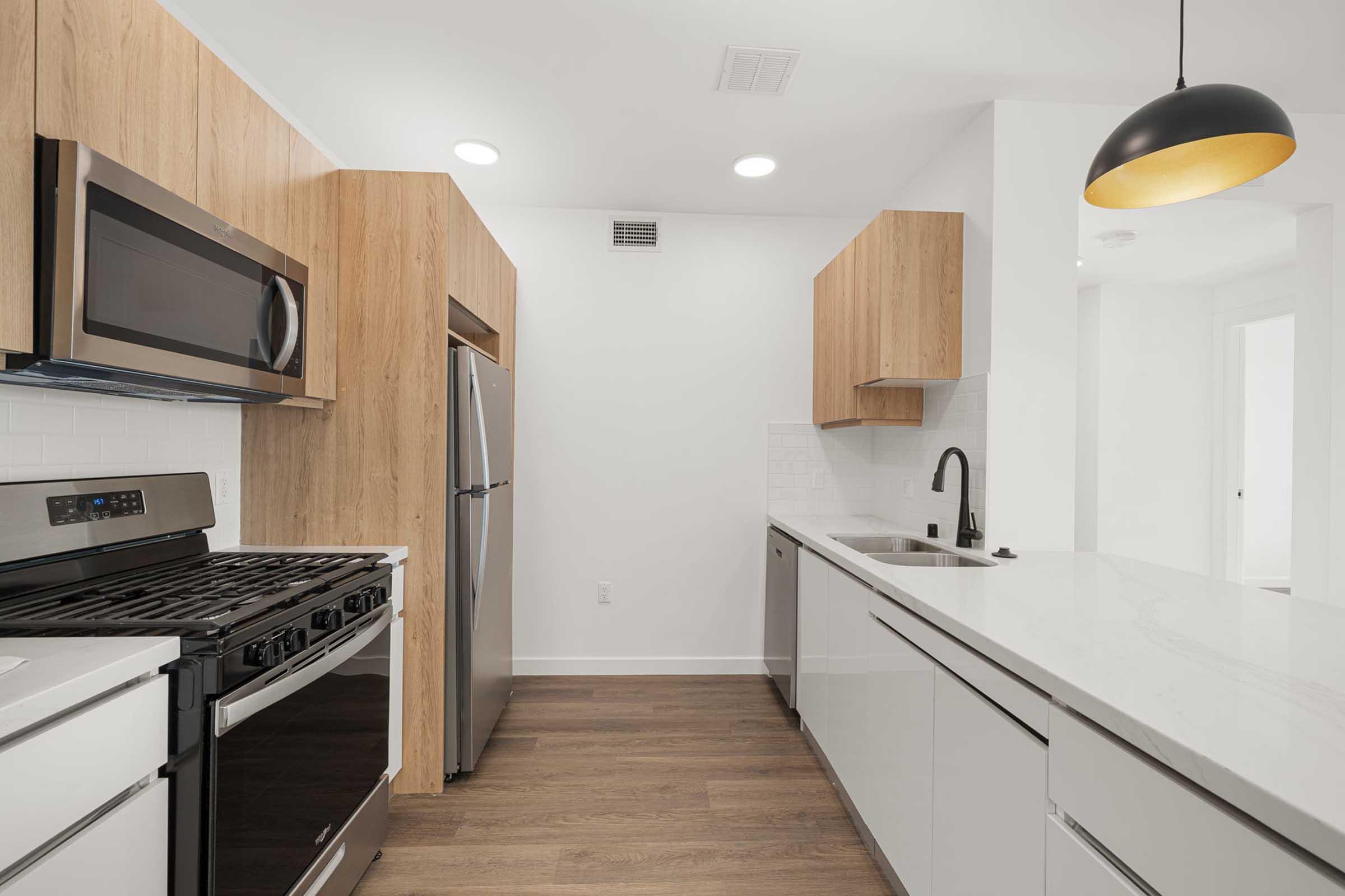 Modern kitchen featuring stainless steel appliances, including a microwave, oven, and refrigerator. The cabinetry has a sleek design with wood accents. A black faucet complements the white countertop, and pendant lighting hangs above the kitchen island. The flooring is a warm wood tone, creating a stylish and inviting space.