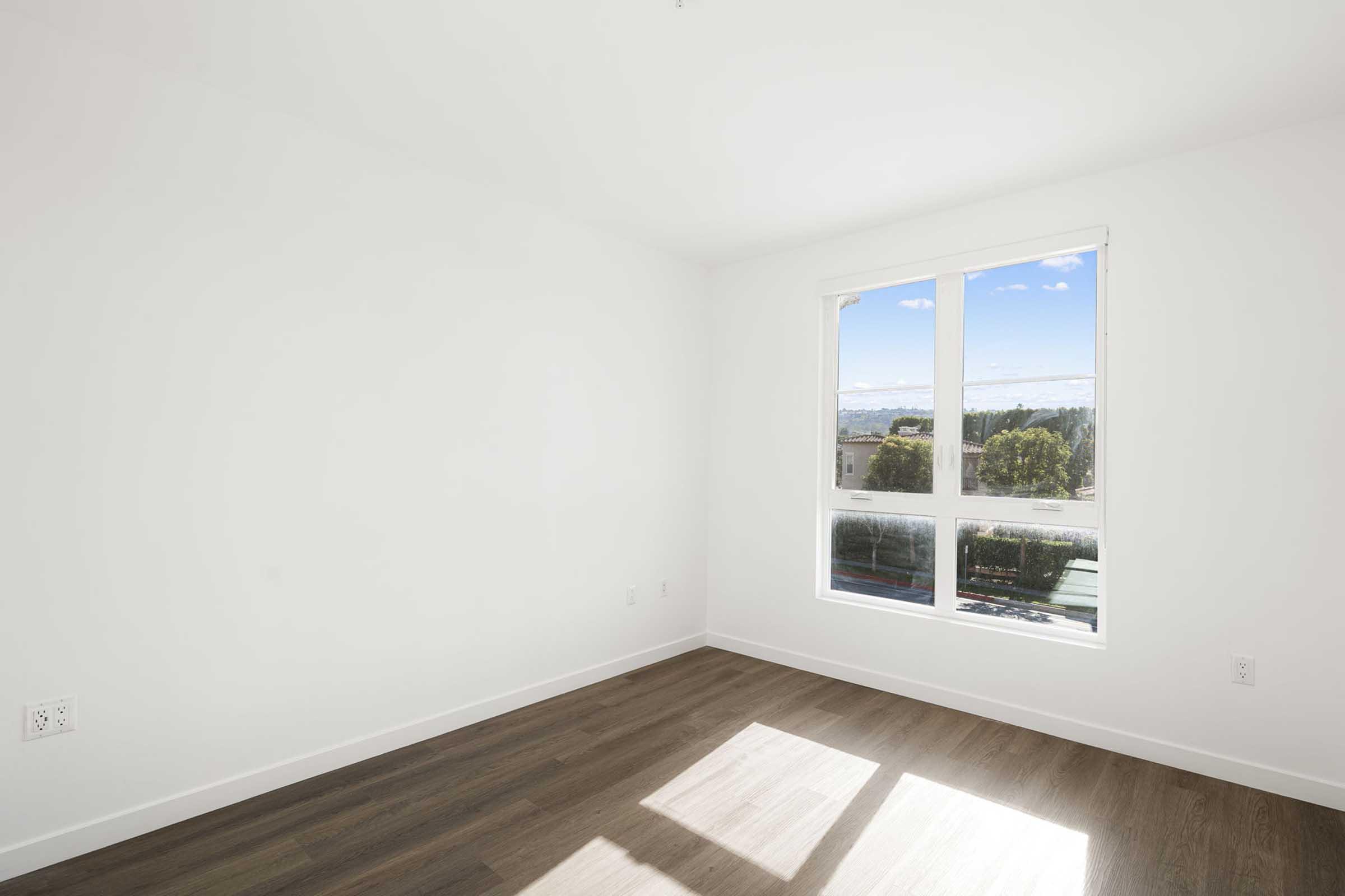 A bright, empty room with white walls and a large window. The window offers a view of greenery and a clear sky. The floor is made of wood, adding warmth to the space. Natural light fills the room, creating an airy and open atmosphere.