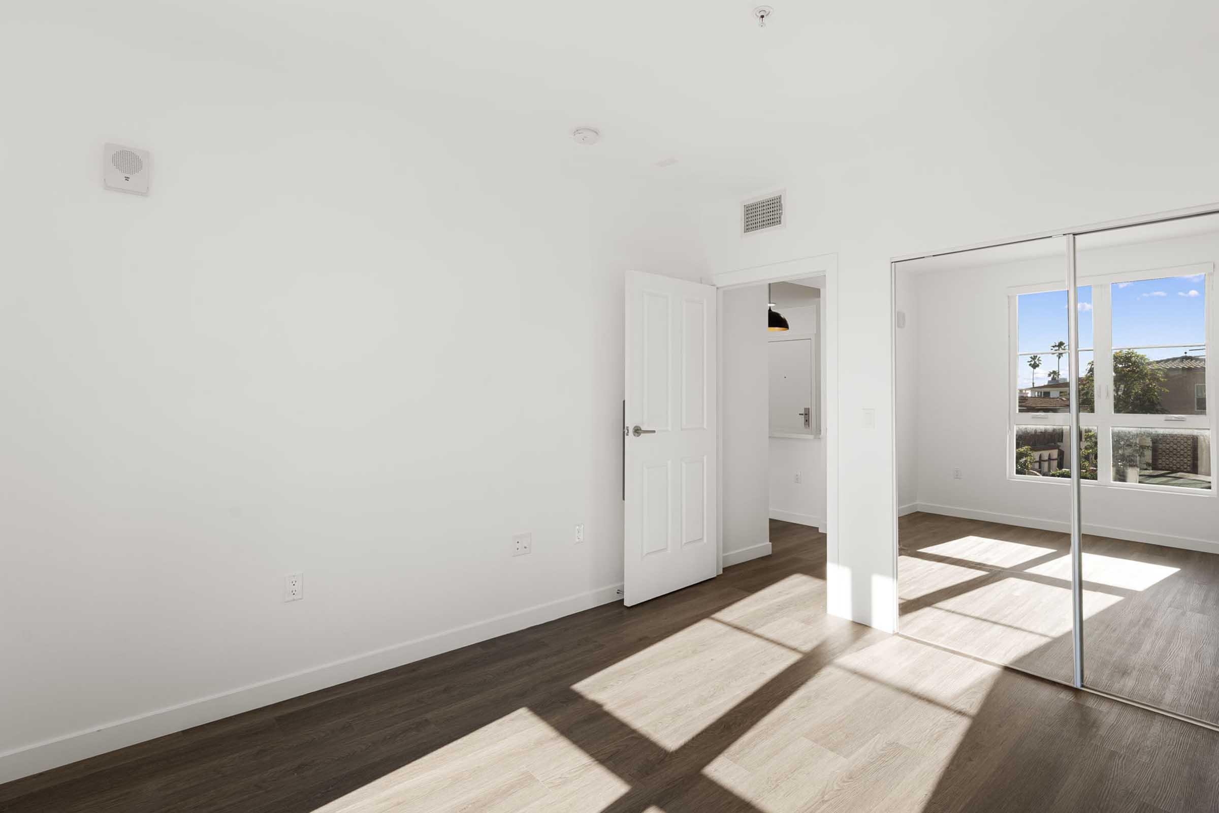 A bright, empty room with white walls and hardwood flooring. A door is partially open, revealing another space beyond. Large windows allow natural light to fill the room, creating a warm atmosphere. There are sliding mirror closet doors on one side.