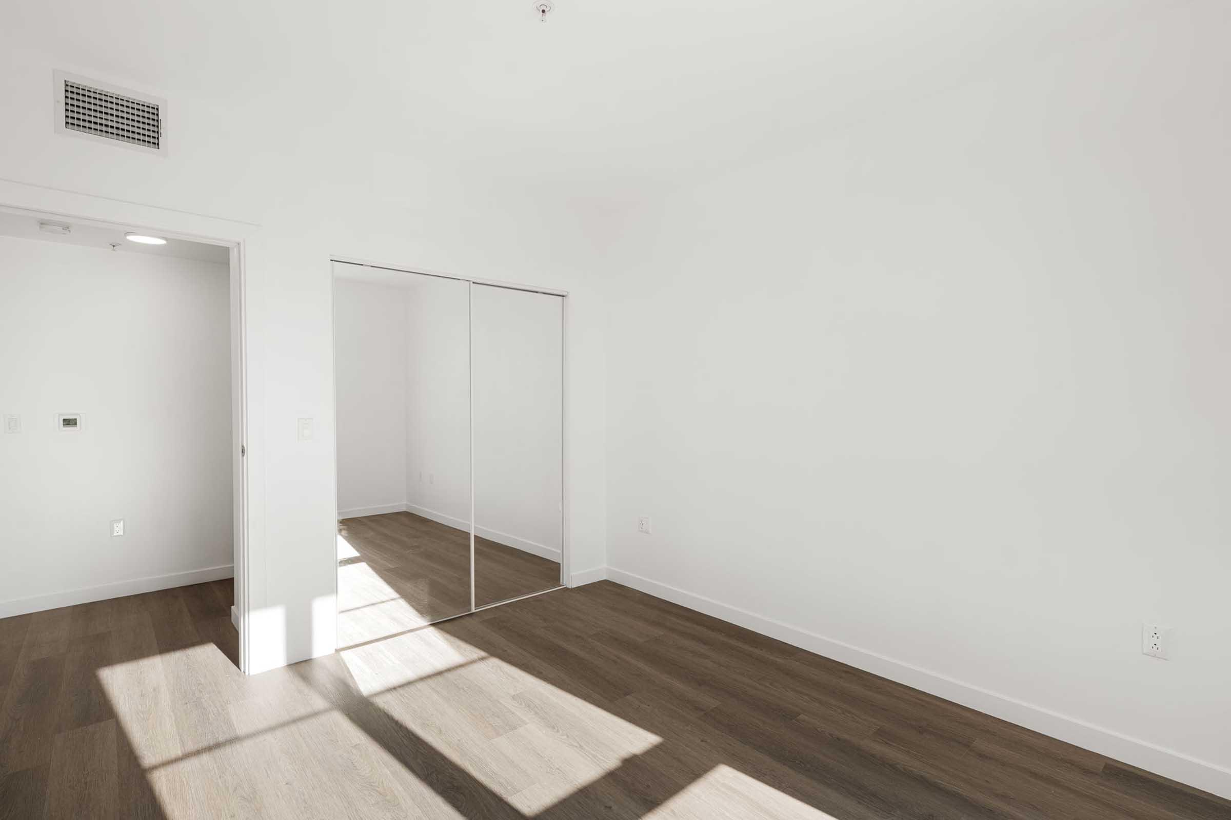 A minimalist room with white walls and a wooden floor. It features a large closet with mirrored doors, allowing natural light to enter through a window, creating a bright and airy atmosphere. The space is empty, ideal for decorating or furnishing as desired.