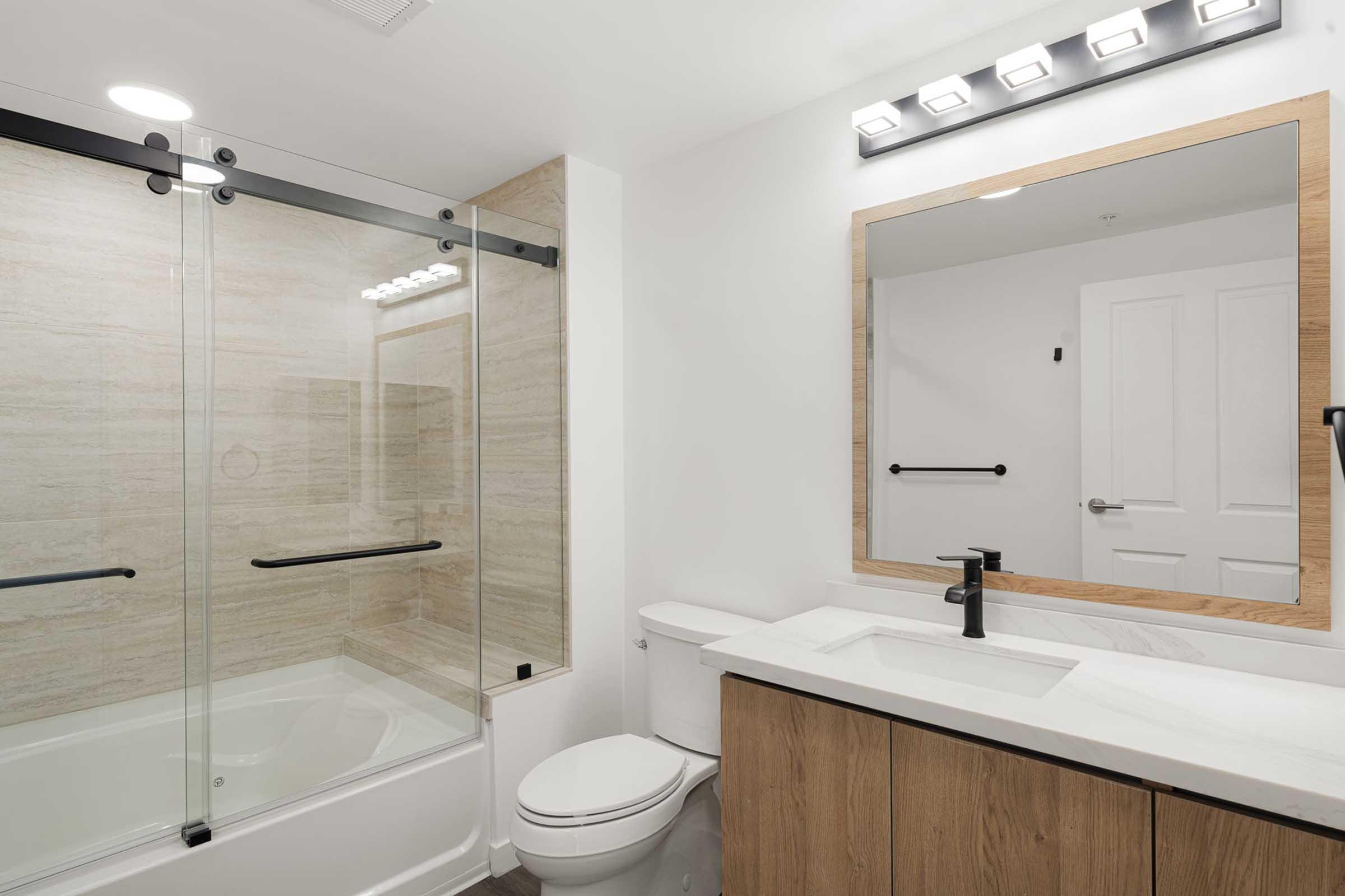 Modern bathroom featuring a glass shower enclosure with sliding doors, a white bathtub, a sleek vanity with a marble countertop, a large mirror, and contemporary lighting. The color scheme includes neutral tones with wooden accents, creating a clean and inviting atmosphere.