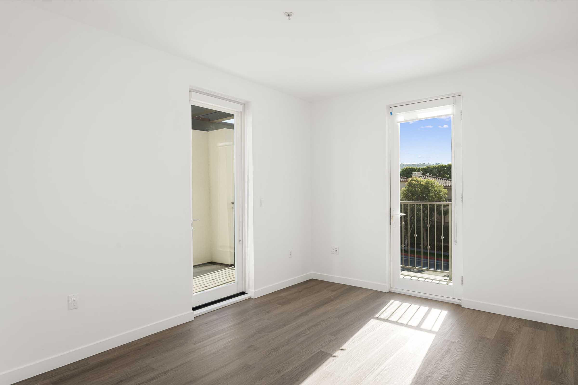 A bright and empty room featuring white walls, two large windows allowing natural light, and wooden flooring. One window opens to a balcony, and the room is spacious with a minimalistic design. The view outside includes greenery and clear blue skies.