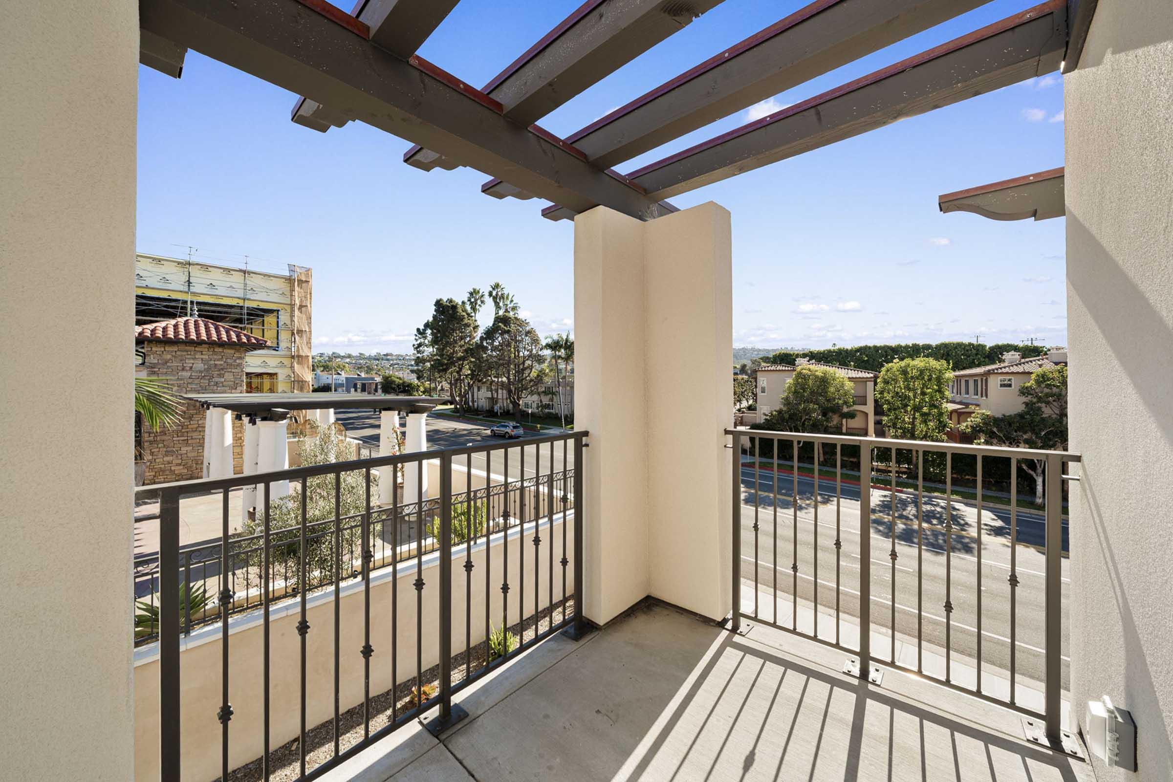 A view from a balcony featuring a railing and overhead pergola structure. The scene includes nearby buildings and trees, with a clear blue sky and scattered clouds above. The perspective overlooks a street, creating an inviting outdoor space.