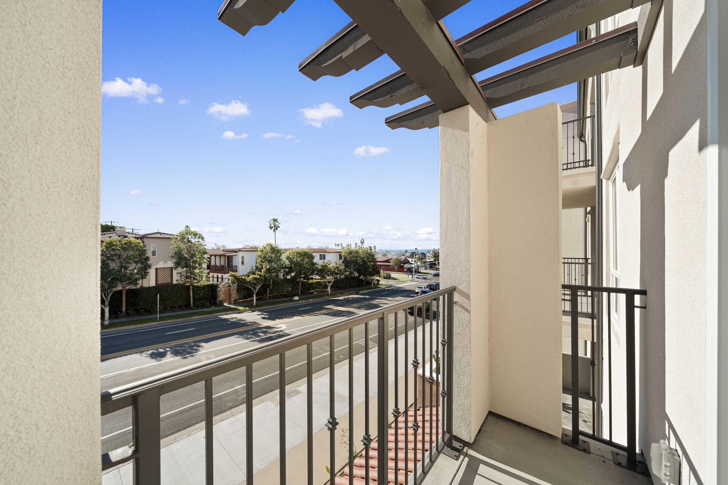 View from a balcony overlooking a street with palm trees and residential buildings. The sky is bright blue with a few fluffy clouds, and the railing creates a modern architectural detail.