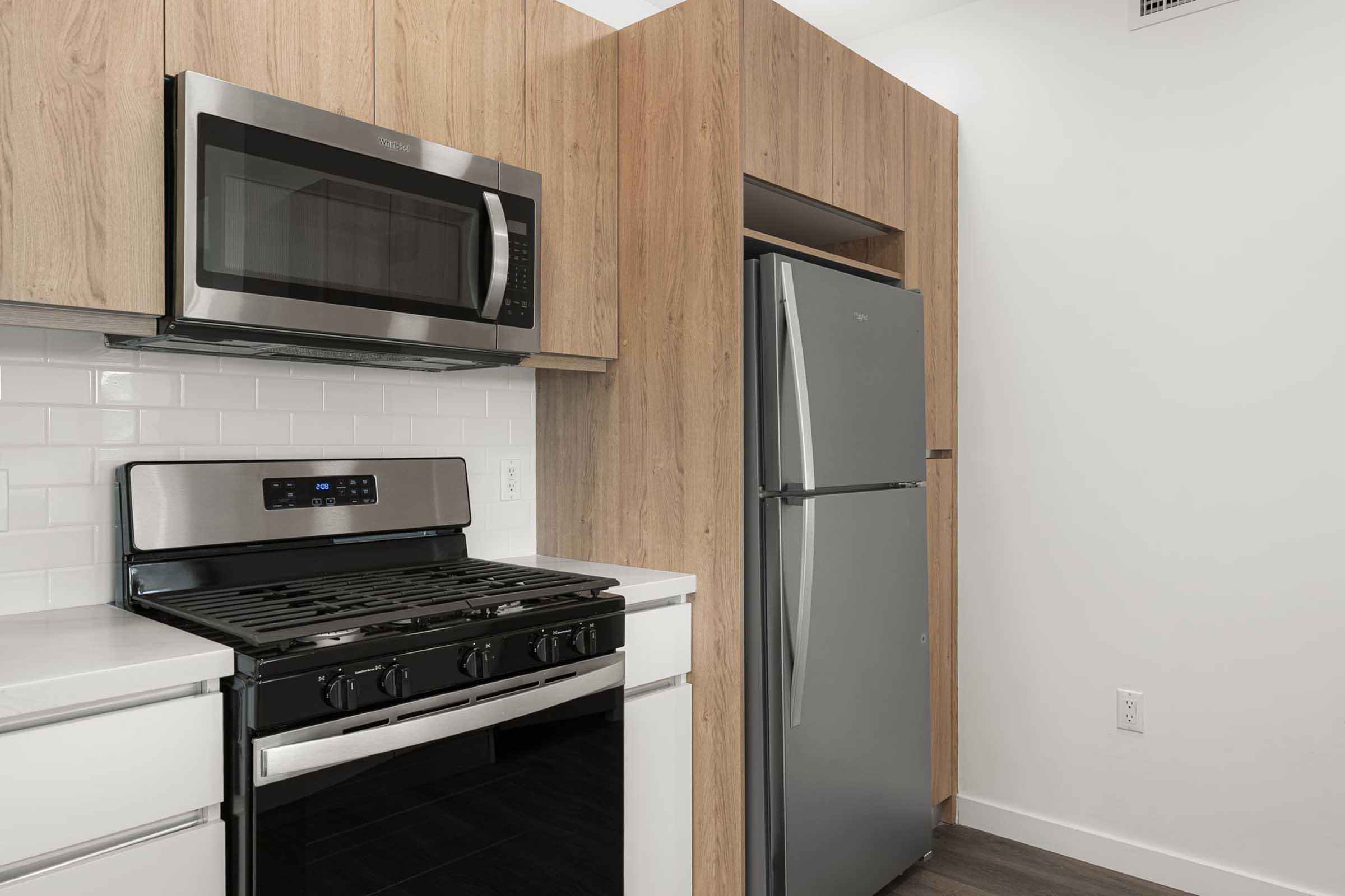 Modern kitchen featuring a stainless steel microwave mounted above a gas stove with a sleek black finish. Adjacent is a stainless steel refrigerator, all set against a backdrop of light oak cabinetry and white tile walls. The overall design is contemporary and functional.