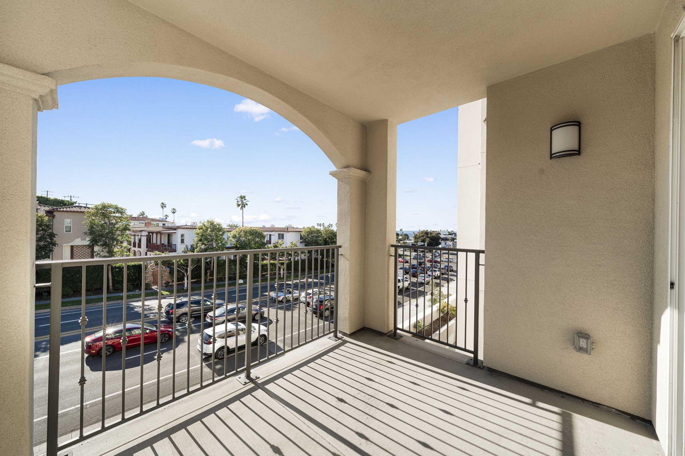 View from a balcony featuring a railing, with a city street below lined with parked cars. The sky is clear with a few clouds, and there are buildings visible in the background, showcasing a suburban setting.