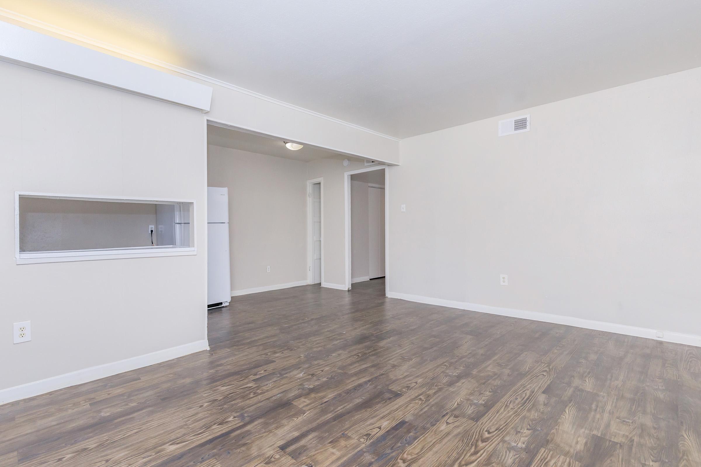 A spacious, well-lit apartment interior with light-colored walls and wood-like flooring. There is an open layout, showcasing a living area leading to a kitchen space with a refrigerator visible. A small serving window is also present, enhancing the connection between the rooms.