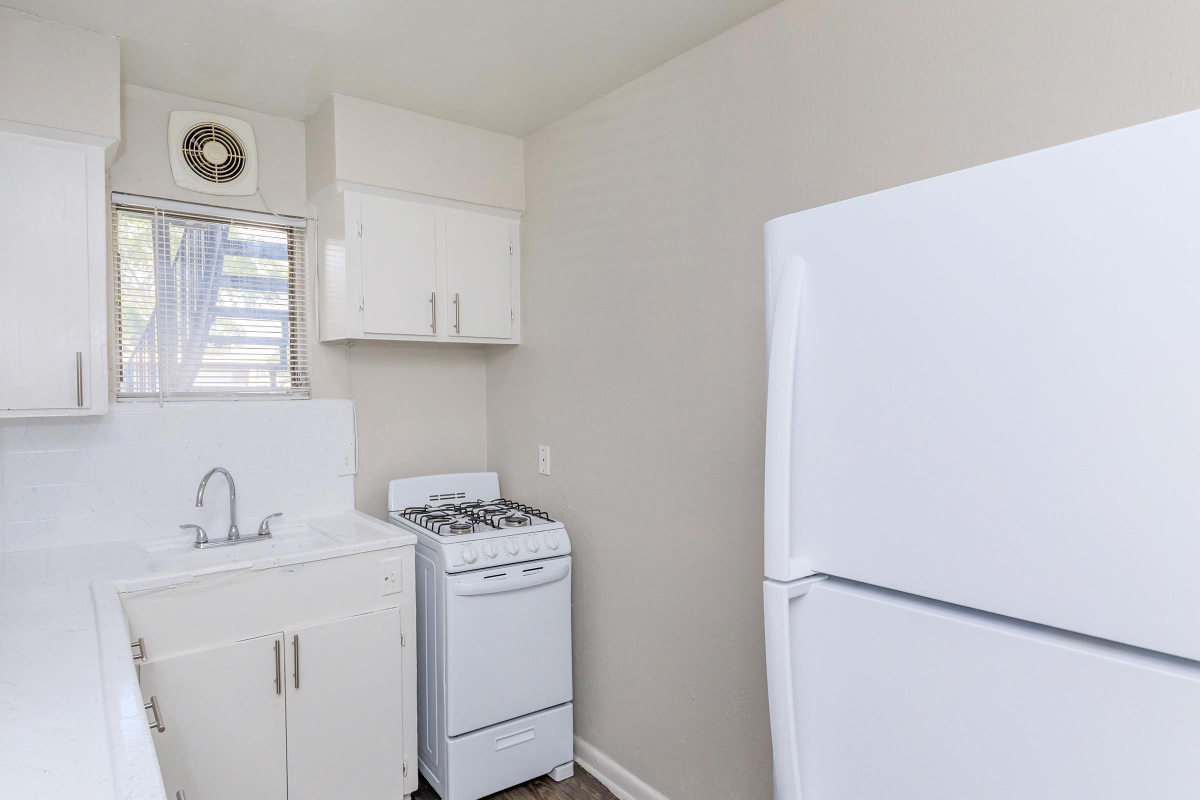 A small, modern kitchen featuring a white refrigerator and a compact white gas stove. The sink is integrated into a white countertop. The walls are painted in a light color, and there is a window with blinds providing natural light. An air vent is visible on the wall above the sink.