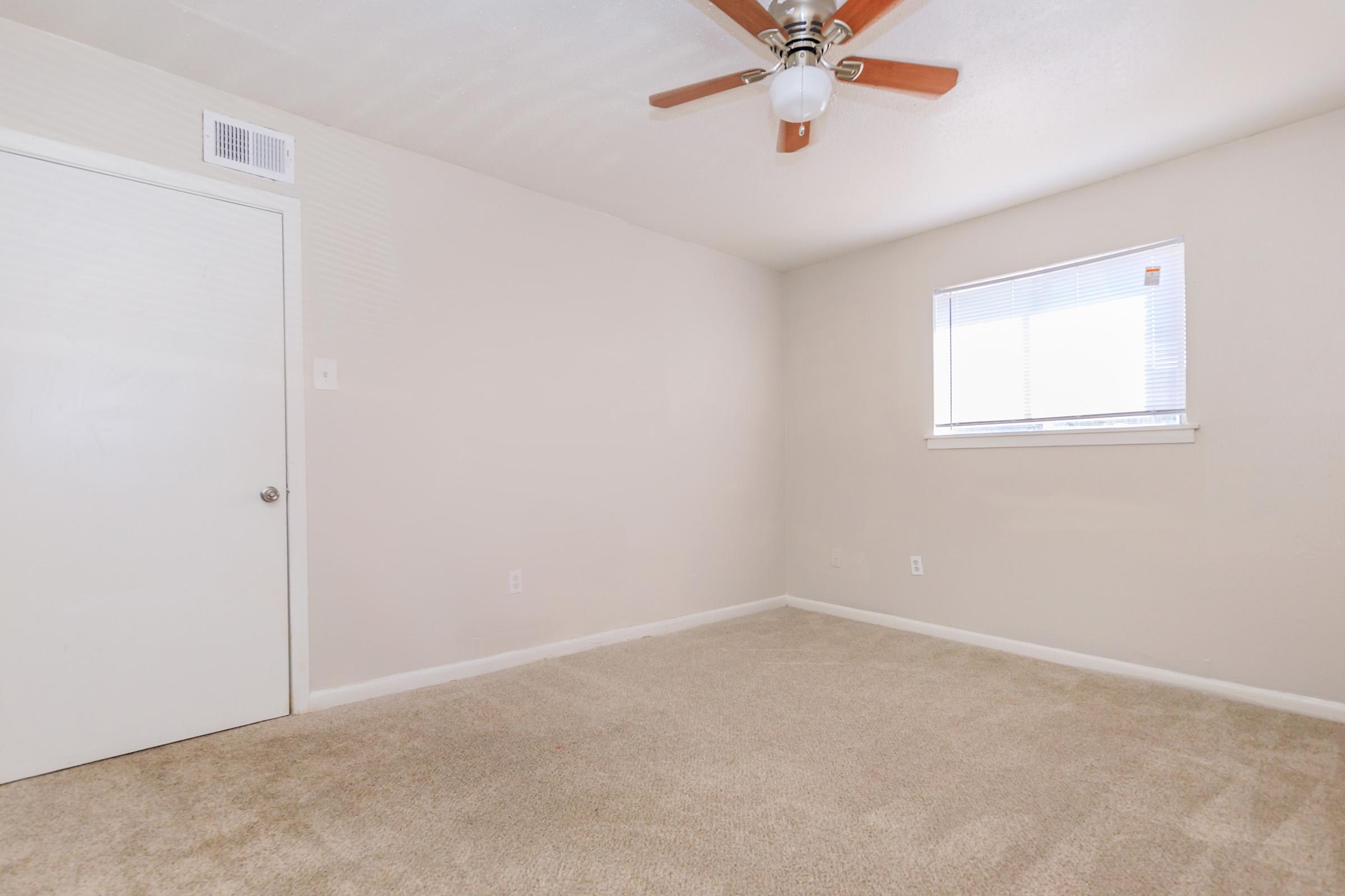 A vacant room with beige walls and carpet. There is a white door on the left and a window on the right, allowing natural light to enter. A ceiling fan with wooden blades is visible. The room appears clean and uncluttered, ideal for various purposes like an office or bedroom.
