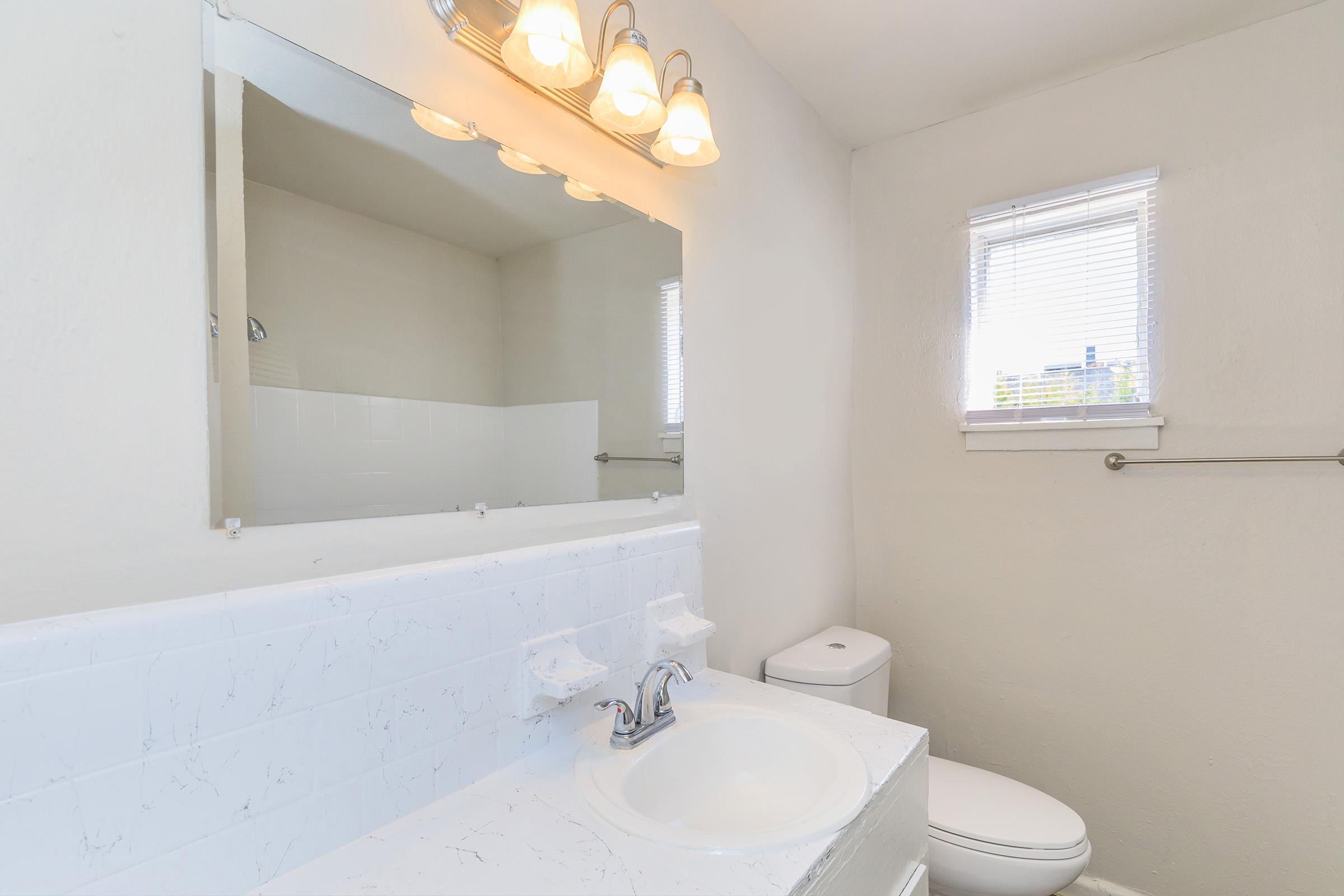A clean and modern bathroom featuring a white countertop with a sink, a large mirror with three light fixtures above, and a window allowing natural light in. The decor includes a white toilet and a simple shower area in the background, with light-colored walls creating a spacious feel.