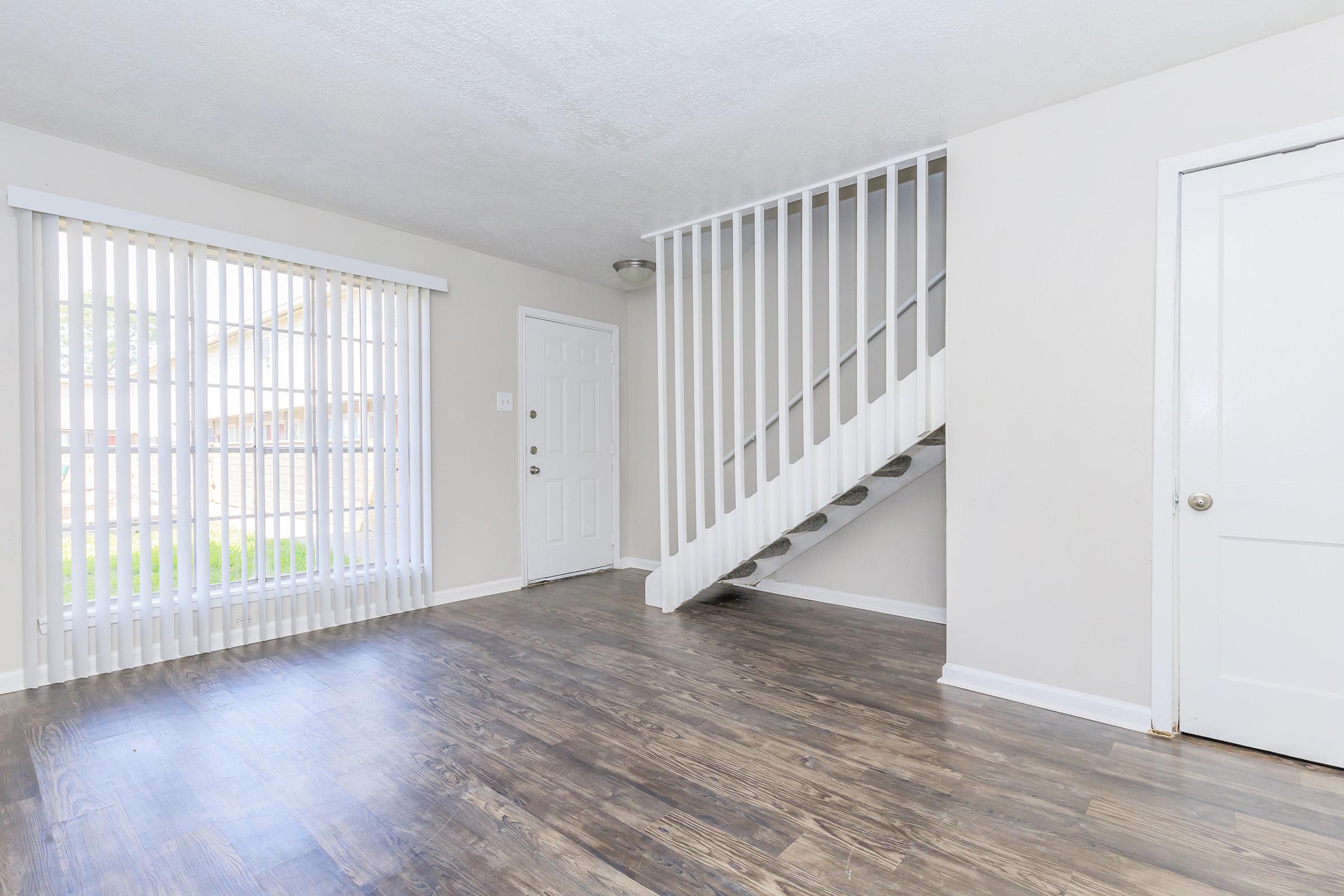Interior view of a spacious living area featuring a staircase, light-colored walls, and a window with vertical blinds allowing natural light. The flooring is a rich, dark wood. A white door leads to the outside, and another door is visible to the right. The space appears clean and well-maintained.