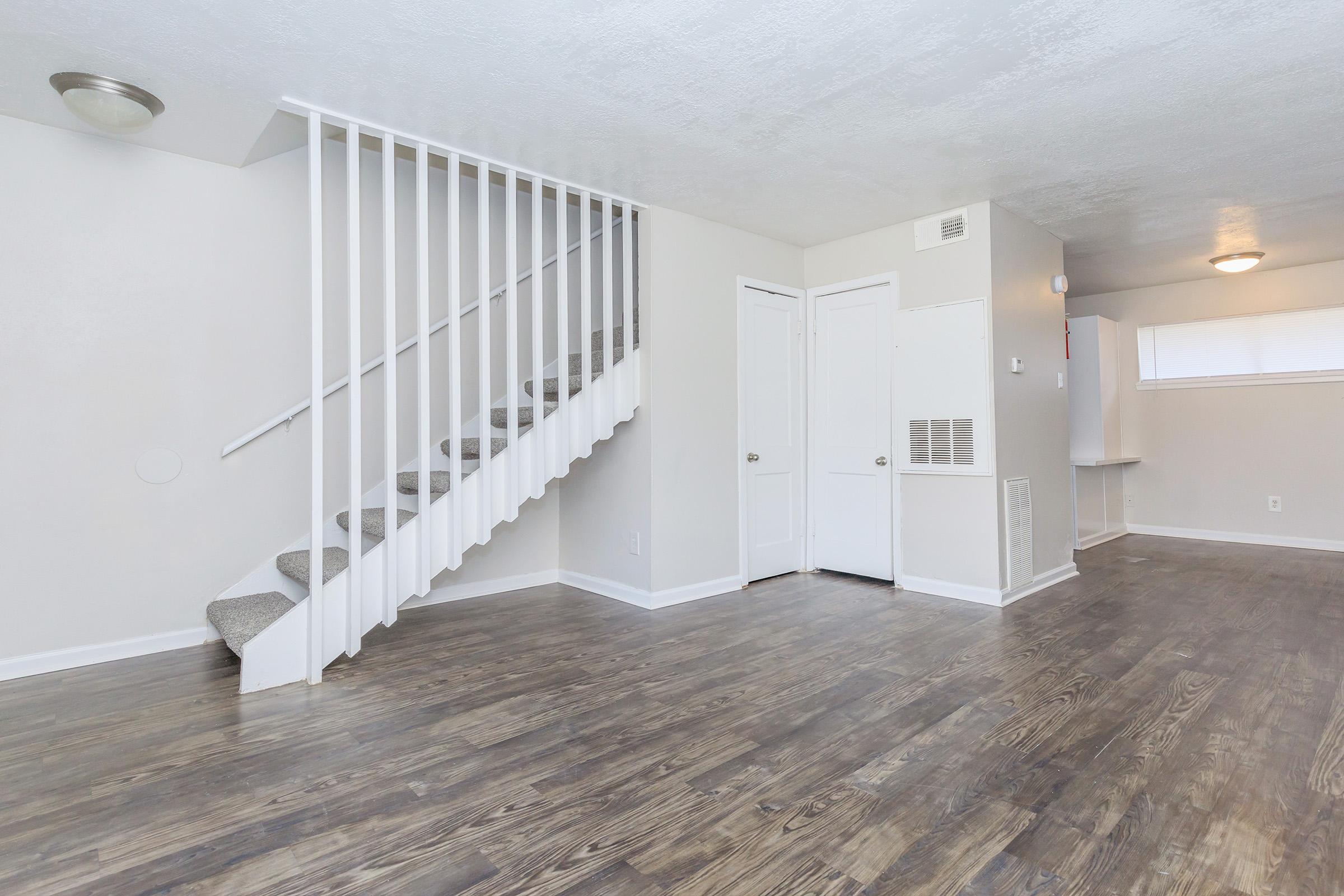 A modern, empty living space featuring light-colored walls and wooden flooring. A staircase with white railing is visible in the background, leading to an upper level. The room has a door on the right and a small kitchen area in the corner, illuminated by natural light from a window.