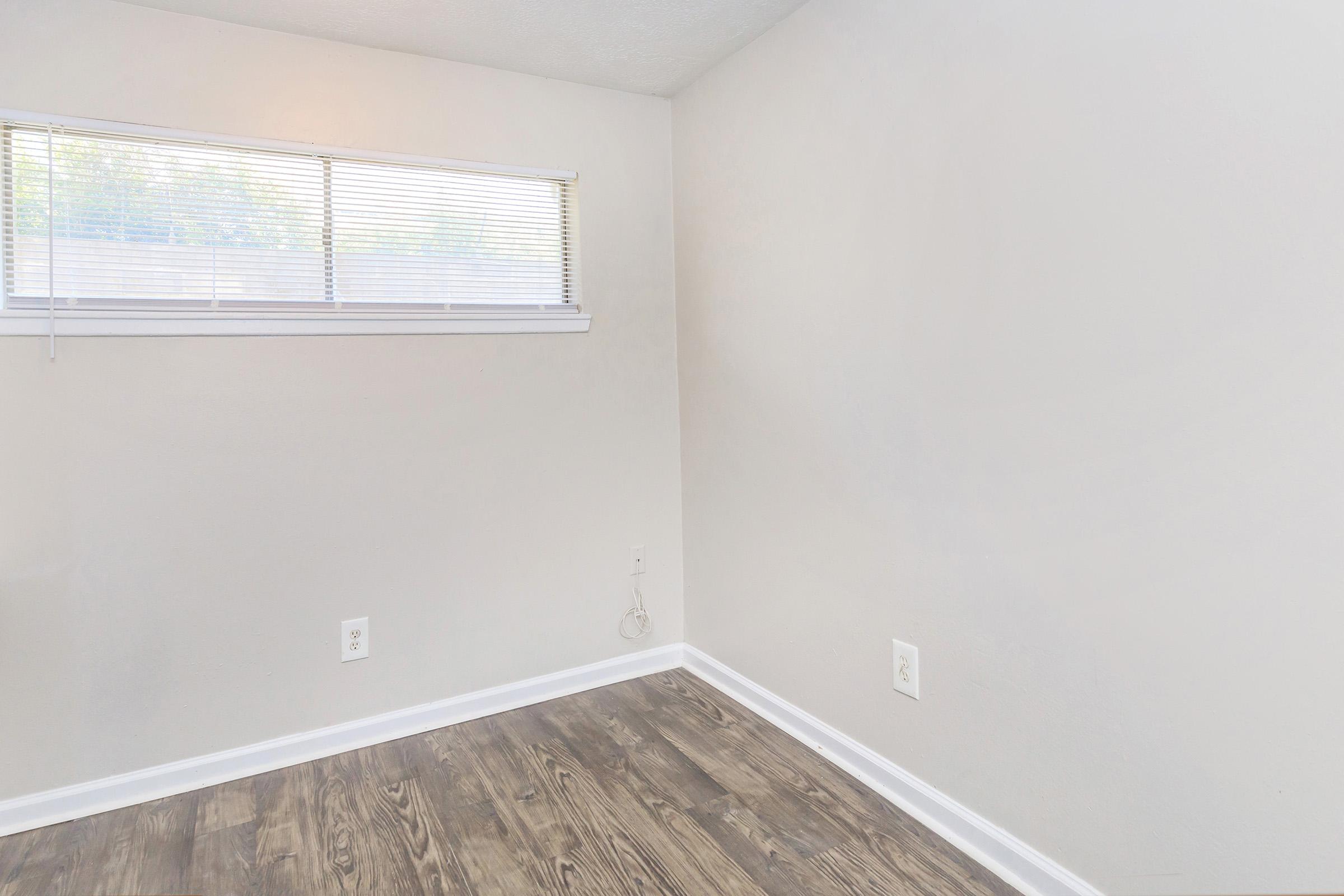 Empty room with light-colored walls and hardwood-style flooring. A small window with white blinds lets in natural light. There are no furnishings or decorations, creating a minimalist and open space.