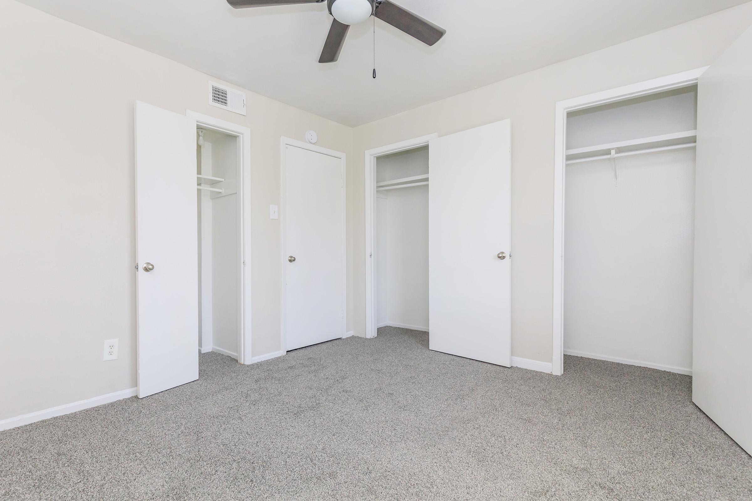 A spacious, empty room with light beige walls and a light gray carpet. It features a ceiling fan and three white doors leading to small closets, with one closet visible on the left side and two on the right. Natural light brightens the space, giving it a clean and airy feel.