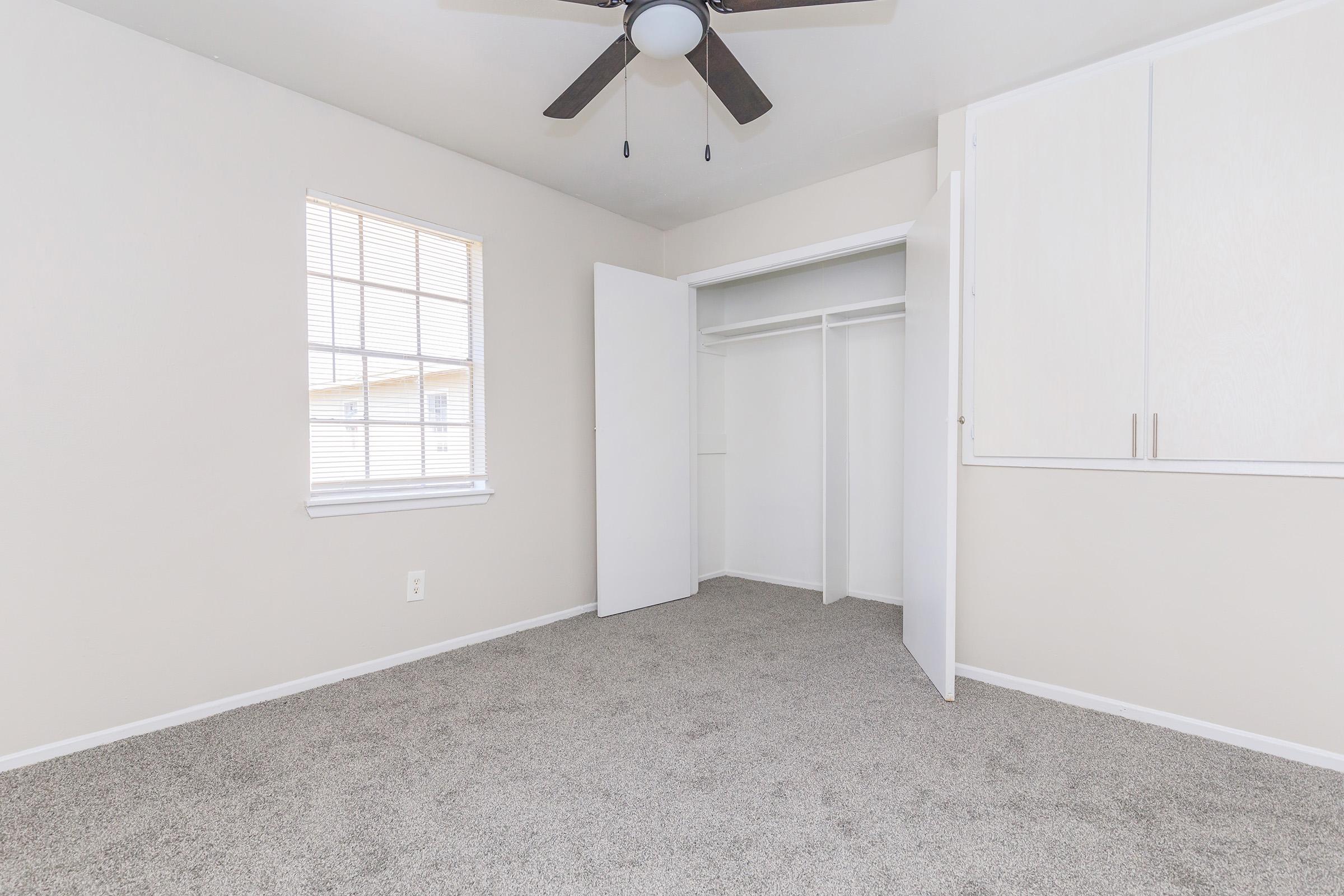 An empty, well-lit bedroom with light-colored walls and carpeted flooring. There is a ceiling fan with dark blades, a window with multiple panes that brings in natural light, and a closet with sliding doors. The overall atmosphere is clean and spacious, ready for furnishing.