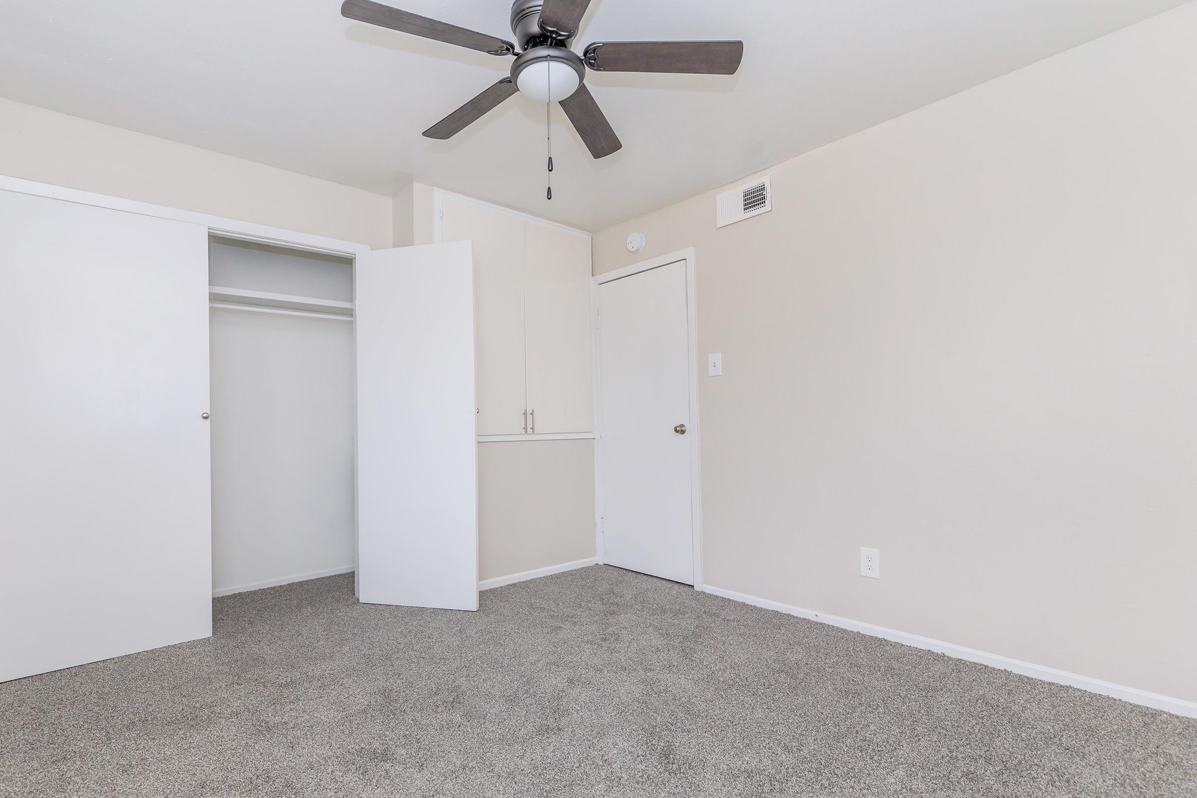A vacant room featuring light beige walls, a ceiling fan, and grey carpet. There are two white doors; one is an open closet and the other leads to a hallway. The room is well-lit and spacious, creating a simple and clean aesthetic.
