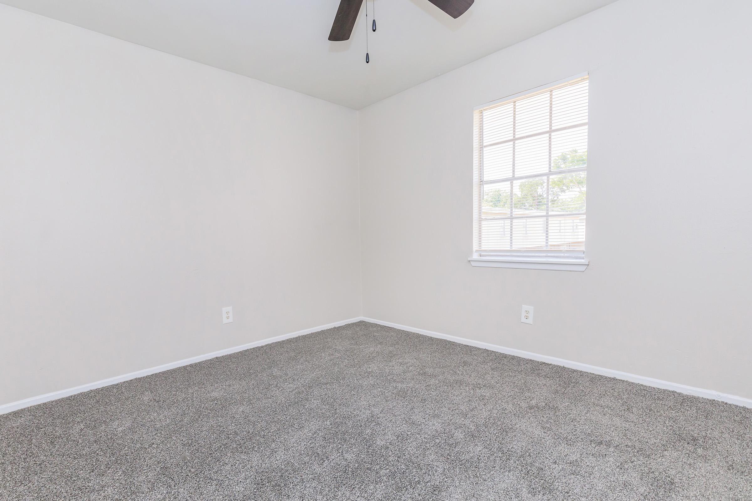 Empty room with light-colored walls and a ceiling fan. The floor is covered with gray carpet, and there is a window with multiple panes allowing natural light to enter. The overall atmosphere is bright and spacious, suitable for various uses.