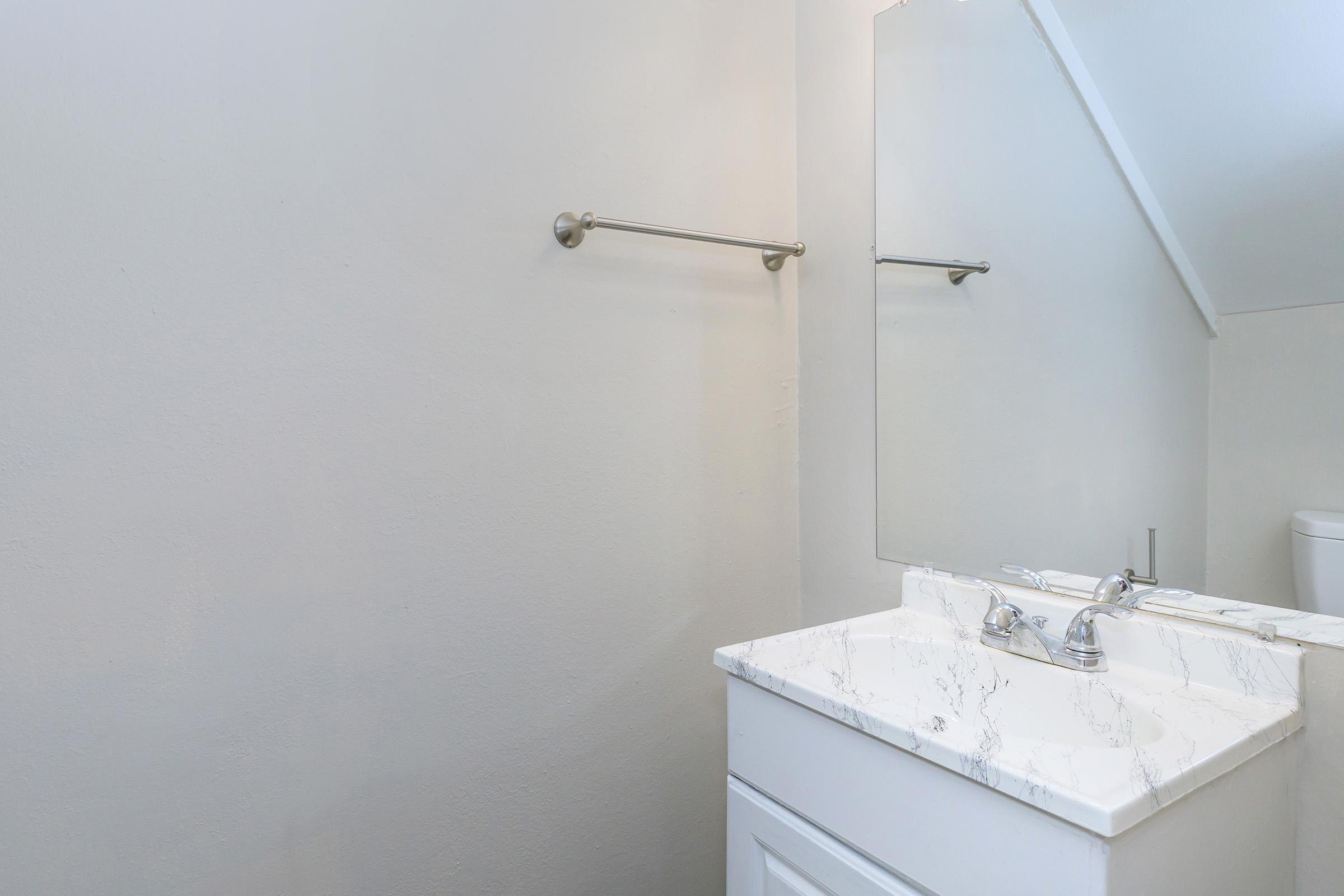 A clean, minimalistic bathroom featuring a white sink with a marble countertop, silver faucets, and a wall-mounted mirror. The walls are painted in a light color, and the space is well-lit, enhancing its simple and modern design. A glimpse of a toilet can be seen in the background.