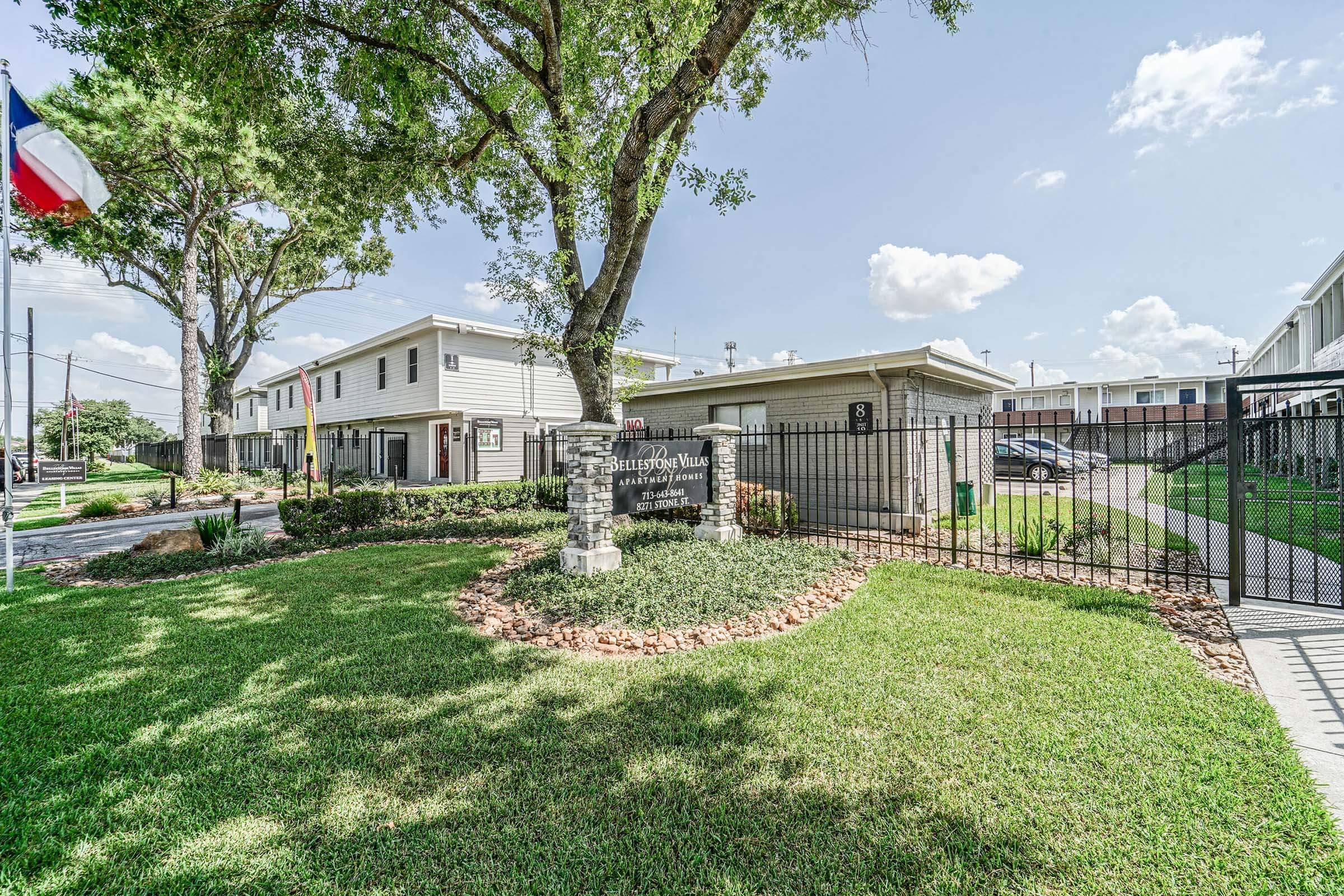 A well-maintained entrance to an apartment complex featuring a sign that reads "Bridges at Vista," surrounded by lush green grass and trees. The entrance is secured with a gate, and there are flags on display. In the background, several buildings are visible under a clear blue sky.