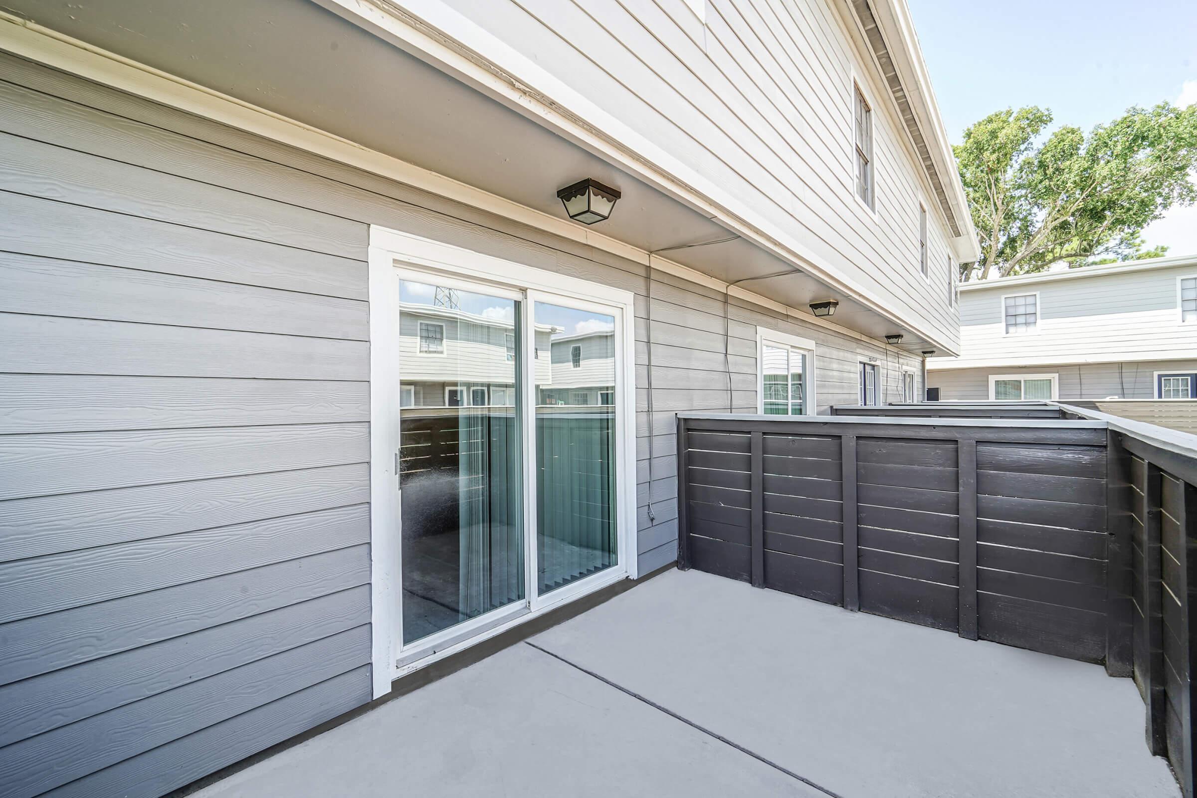 A patio area with a sliding glass door leading to a house, featuring gray siding on the walls. The floor is concrete, and there are black wooden railings. In the background, additional units are visible with windows and similar siding. The setting is bright and well-lit, indicating a sunny day.