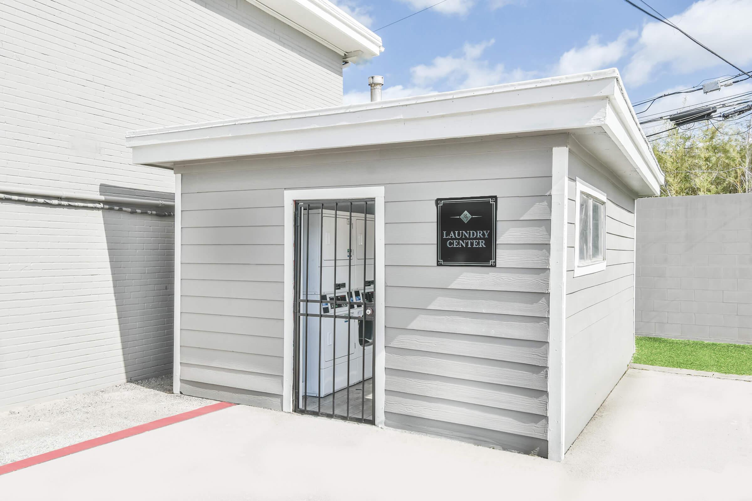 A small gray laundry center building with a sign that reads "LAUNDRY CENTER." The building features a gated entrance, two windows, and is located beside a larger light-colored structure. There are power lines above and a patch of green grass in the background.