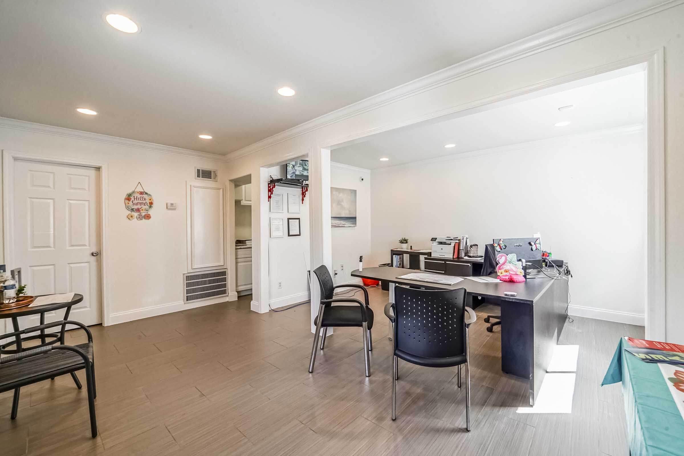 A bright, modern office space featuring a desk with a computer, chairs, and decorative elements on white walls. The floor is tiled in a light color, and natural light enters through bright windows. There is a door leading to another room, enhancing the openness of the area.