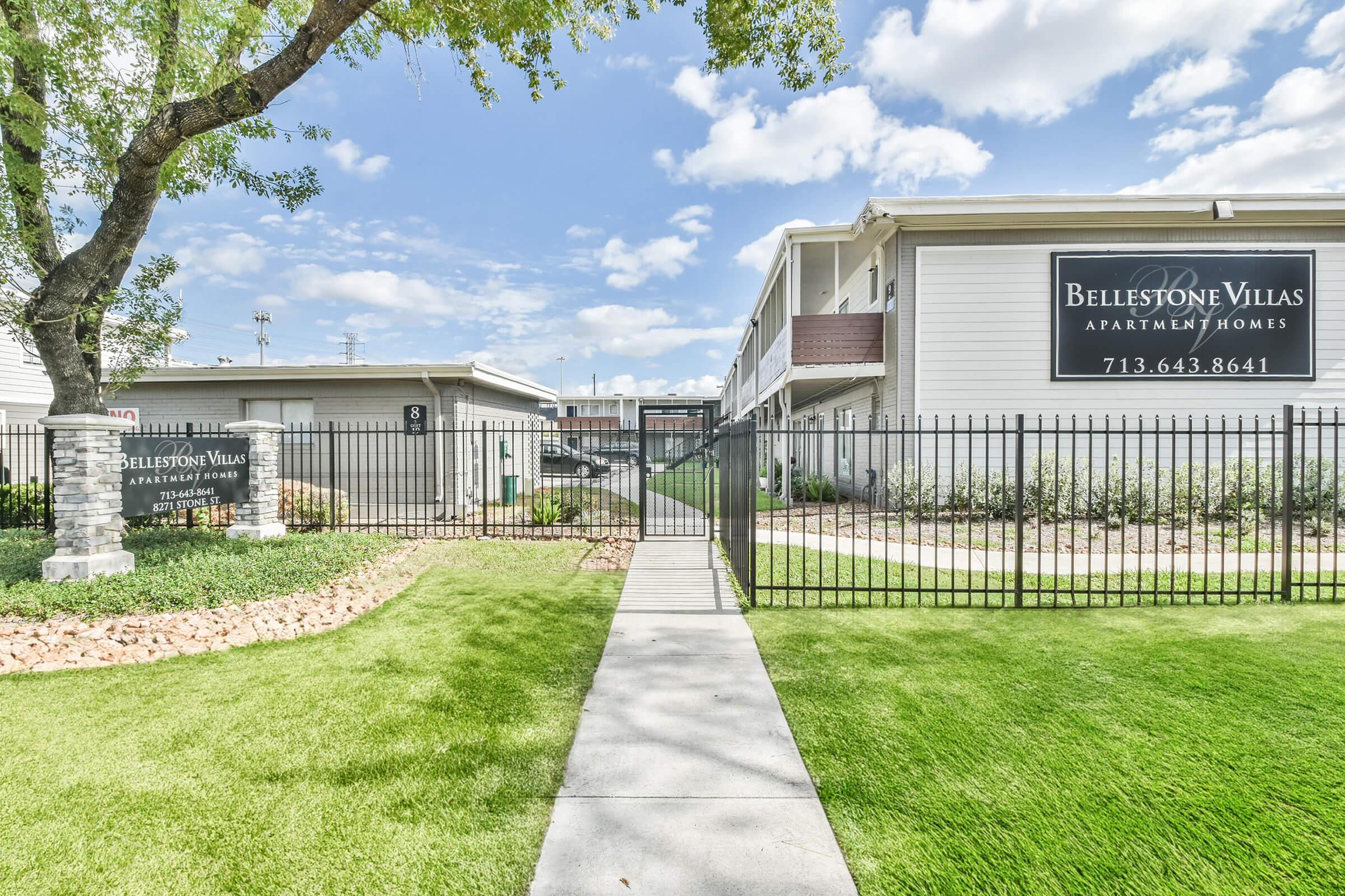 View of the entrance to Bellestone Villas Apartment Homes, featuring a well-maintained pathway leading through a landscaped area with grass and decorative stones. The buildings are visible in the background, and a fence surrounds the complex. Bright blue sky with scattered clouds.