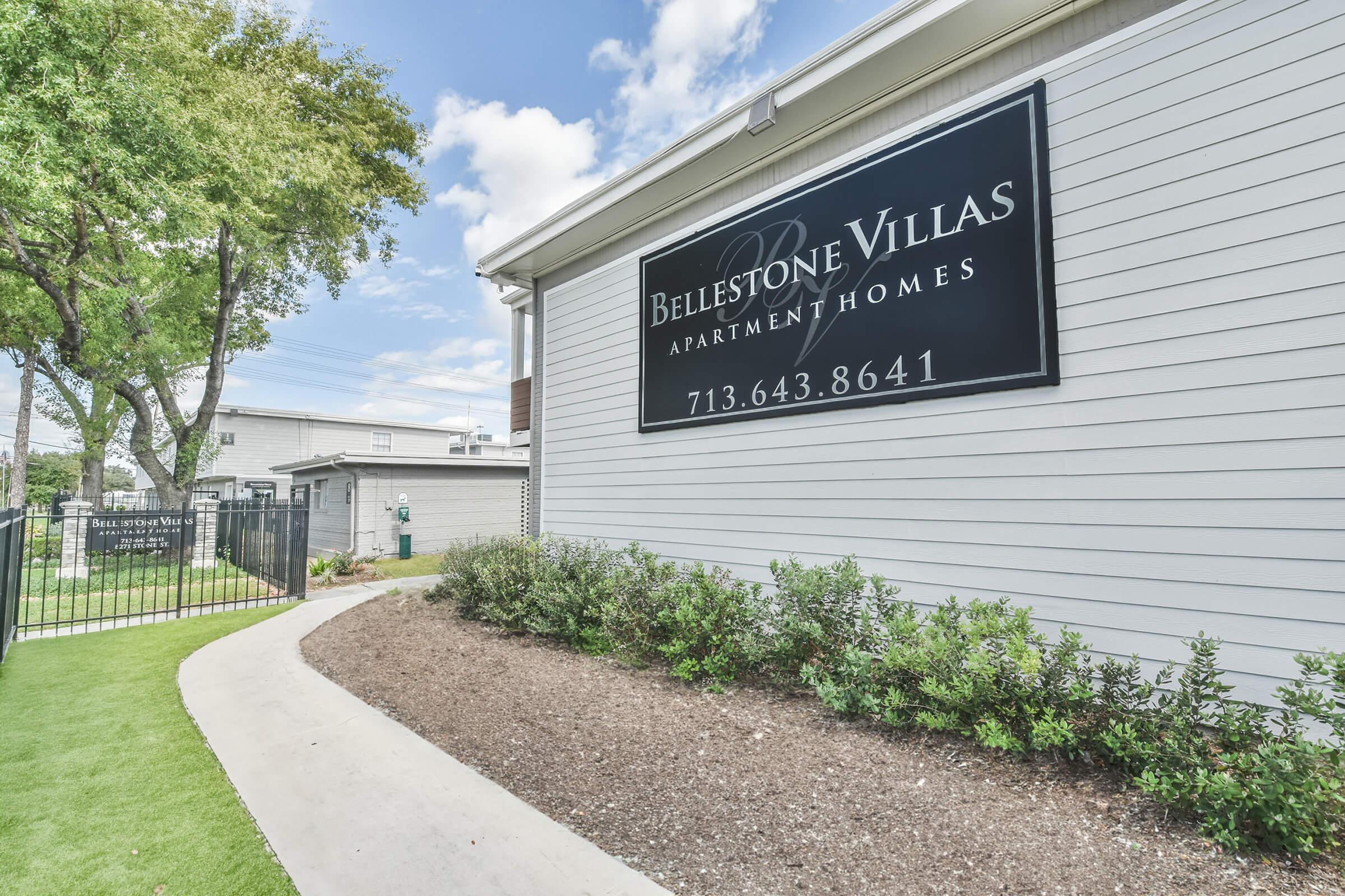Sign for Bellestone Villas Apartment Homes, featuring the name prominently displayed along with a contact number. The sign is set against a light-colored building exterior, with a landscaped pathway leading towards it, surrounded by greenery and trees under a partly cloudy sky.