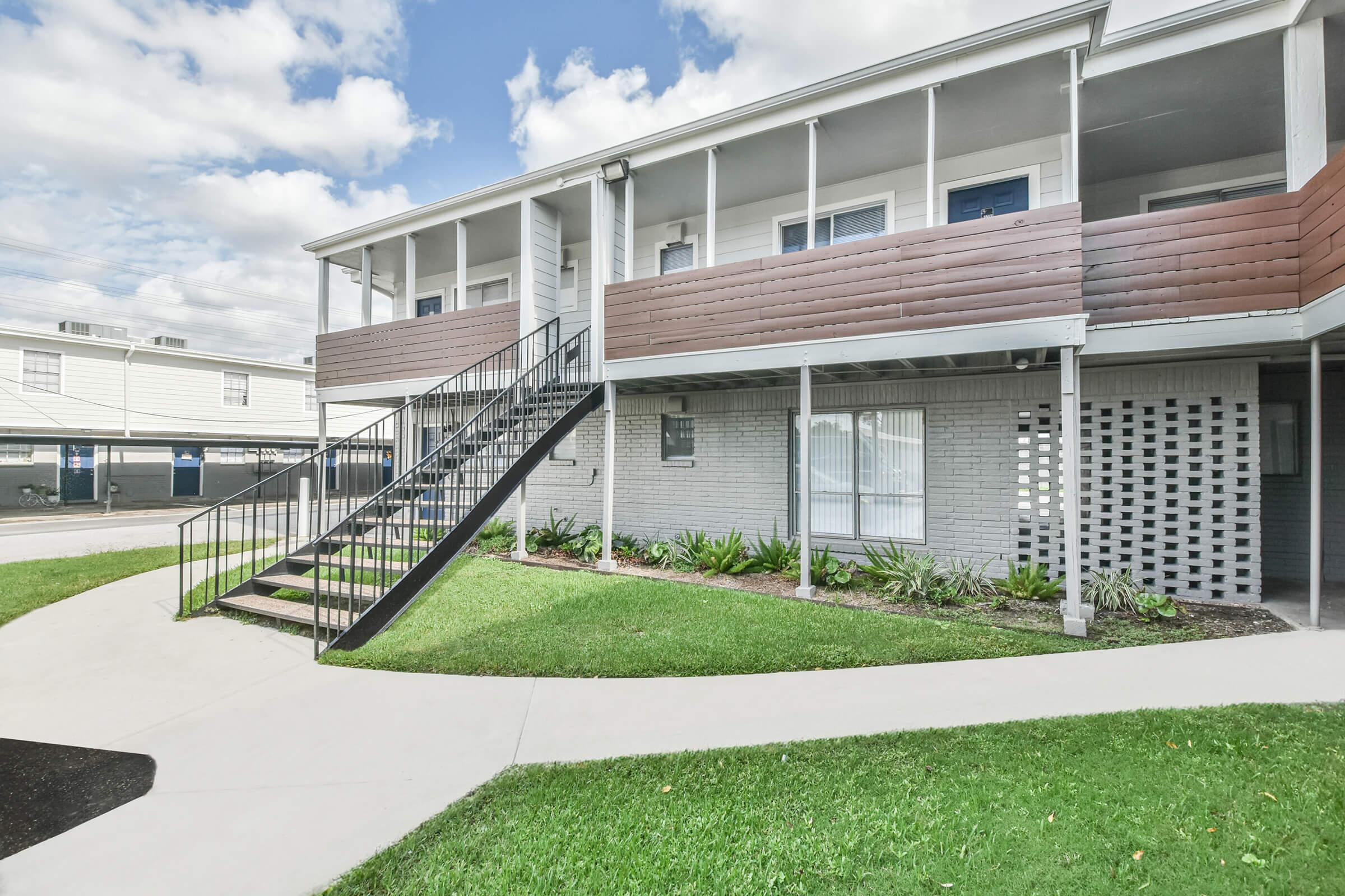 Two-story residential building with a staircase leading to a second-floor entrance. The facade features a mix of light-colored bricks and wooden paneling. Lush green grass and decorative plants surround the building, and there are cloudy blue skies above. Pathways connect different areas of the property.