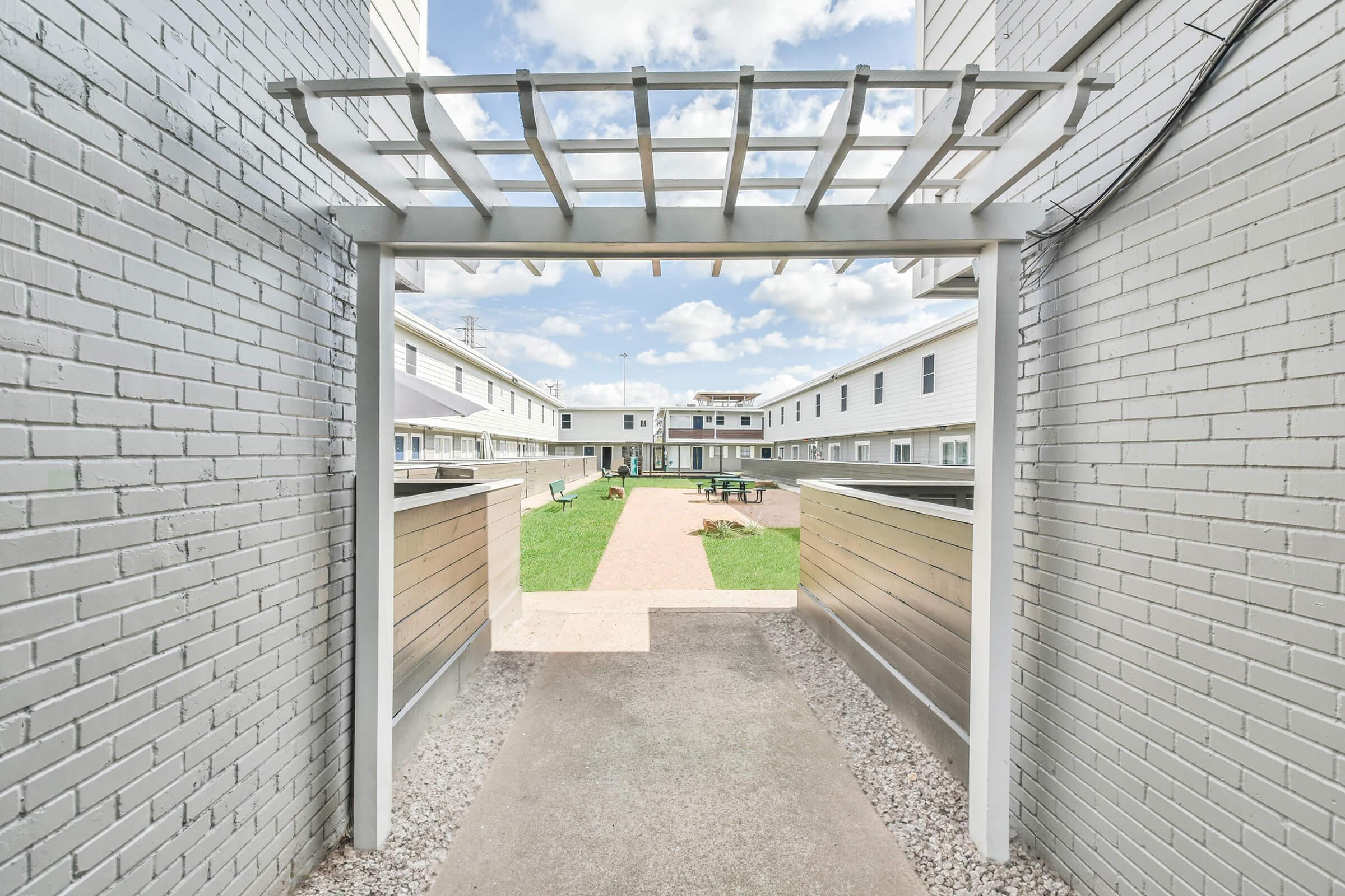 A view through a white trellis archway leading to a grassy courtyard surrounded by two-story buildings. The courtyard features pathways, benches, and scattered trees under a partly cloudy sky.