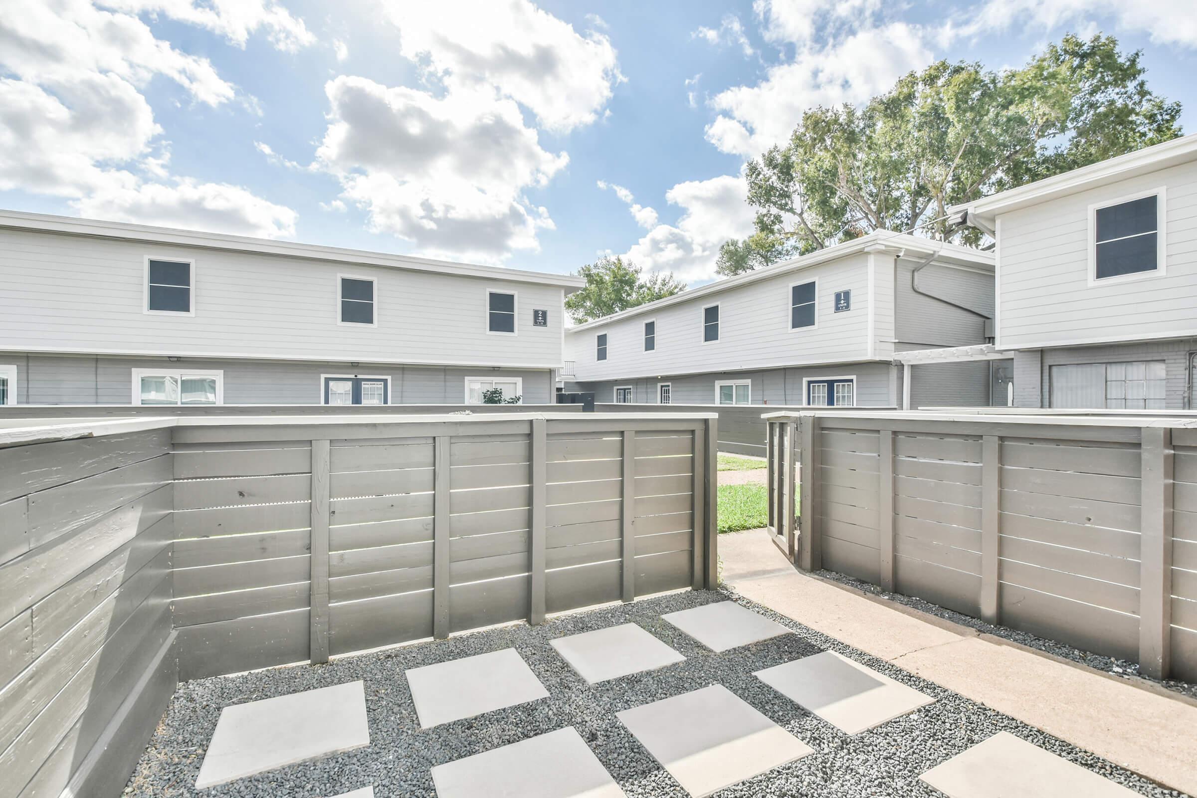A view of a fenced outdoor area with stone pavers and gravel, surrounded by two-story apartment buildings. The scene features a blue sky with scattered clouds and green grass in the background. The layout suggests a communal space within a residential complex.